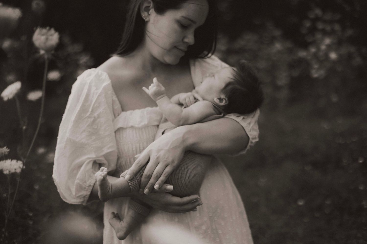 A mother in a white dress tenderly cradles her newborn baby outdoors in a soft sepia-toned portrait.