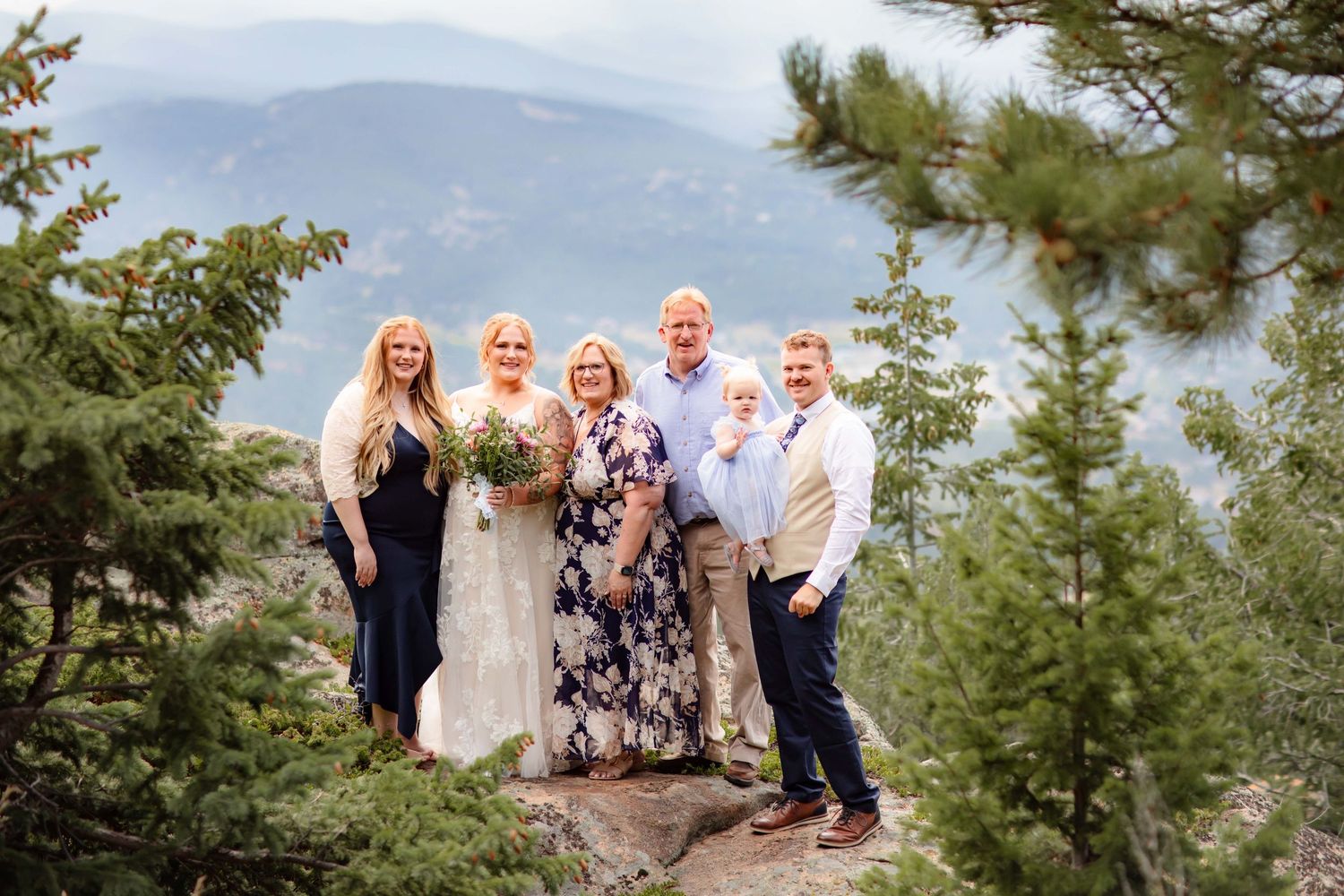 A wedding party poses together on a scenic mountain overlook surrounded by evergreen trees.