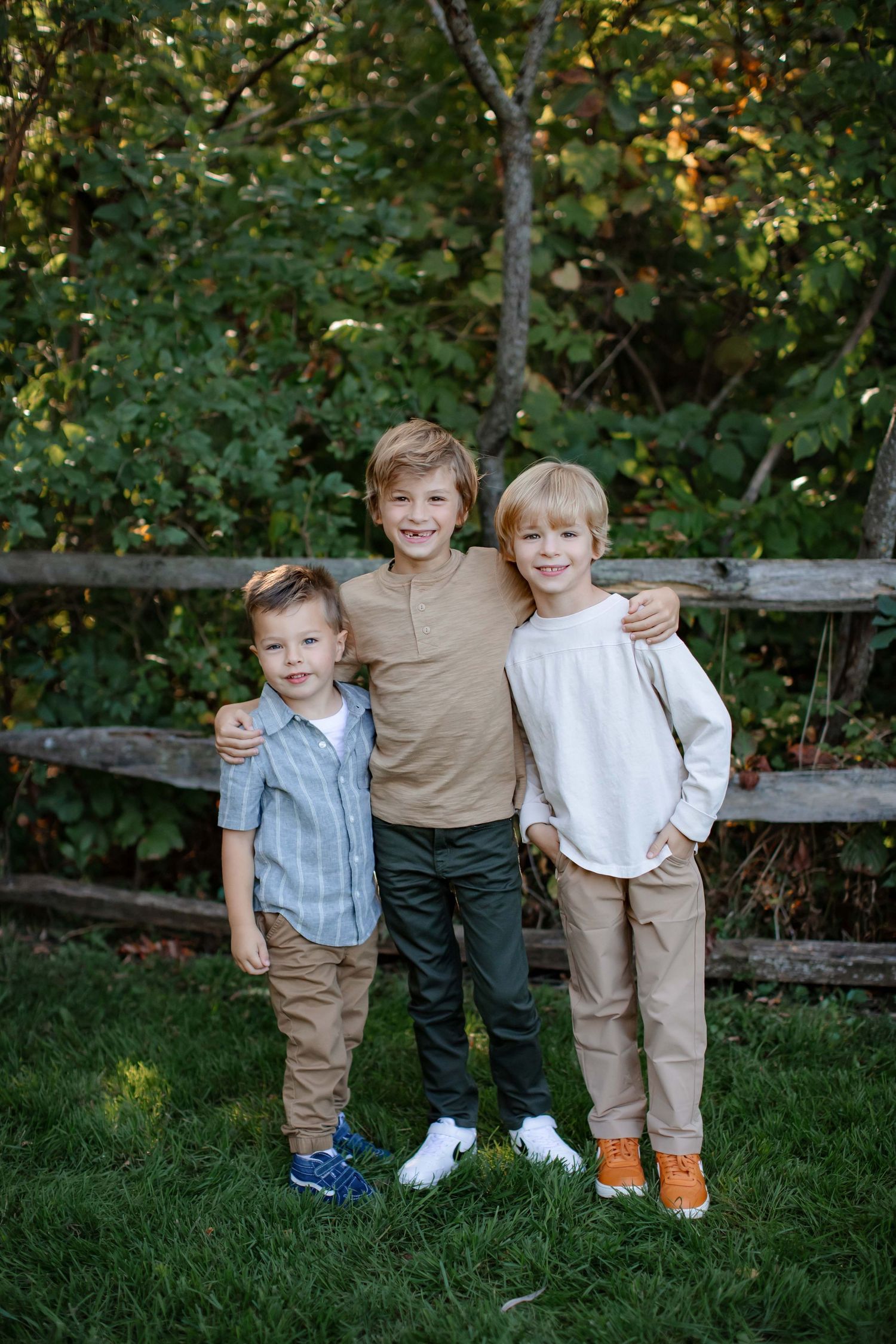 Three young boys in casual clothing pose together in front of wooden fence outdoors.