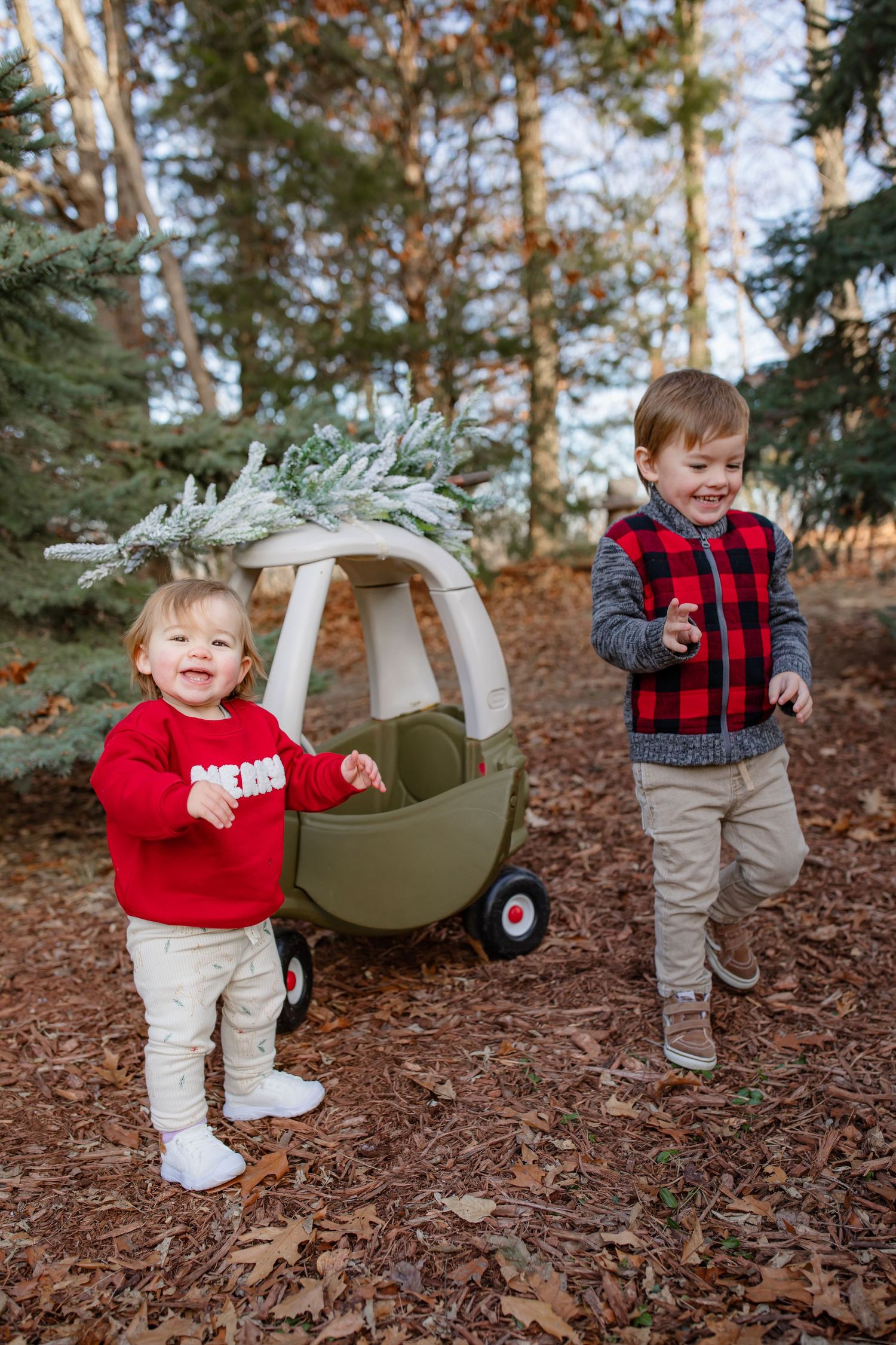 Siblings playing with decorated cozy coupe car on woodland trail during winter photoshoot.
