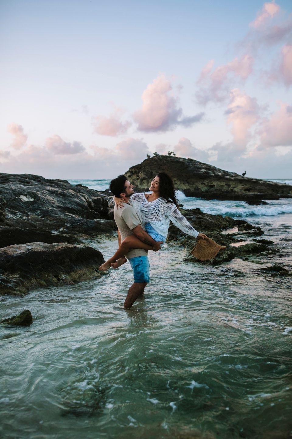 Couple playing in shallow ocean waters near rocky shore during sunset.