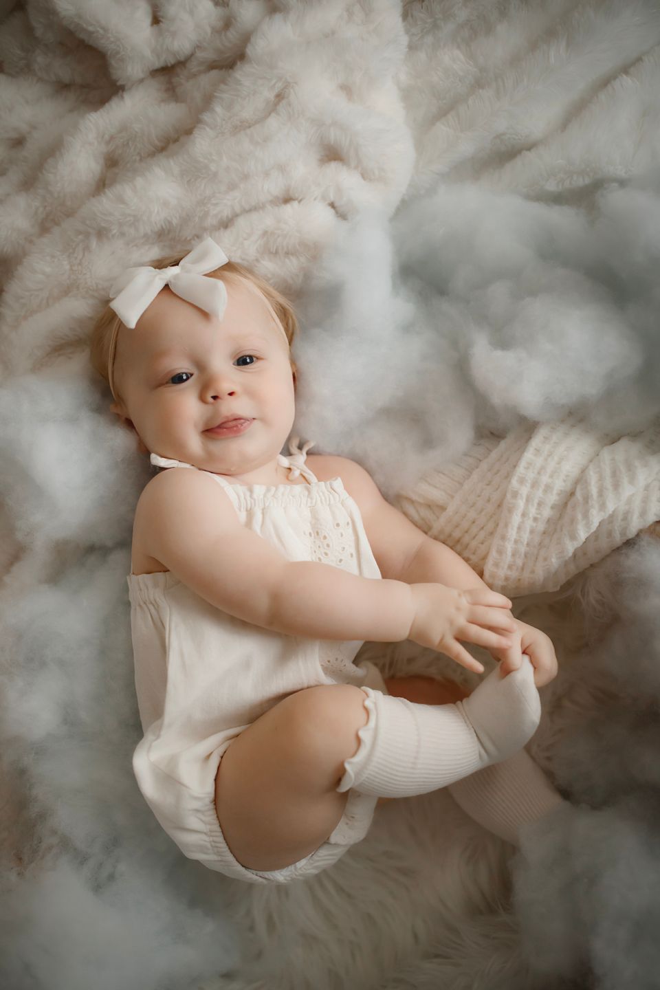 Baby in white outfit sitting among cloud-like textures with gentle smile.