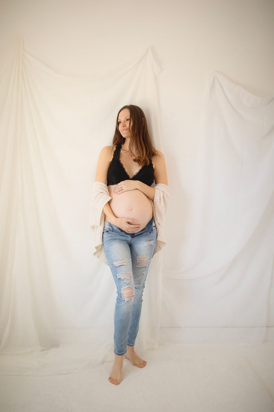 Expectant mother in ripped jeans and black top poses against white backdrop during maternity photoshoot.