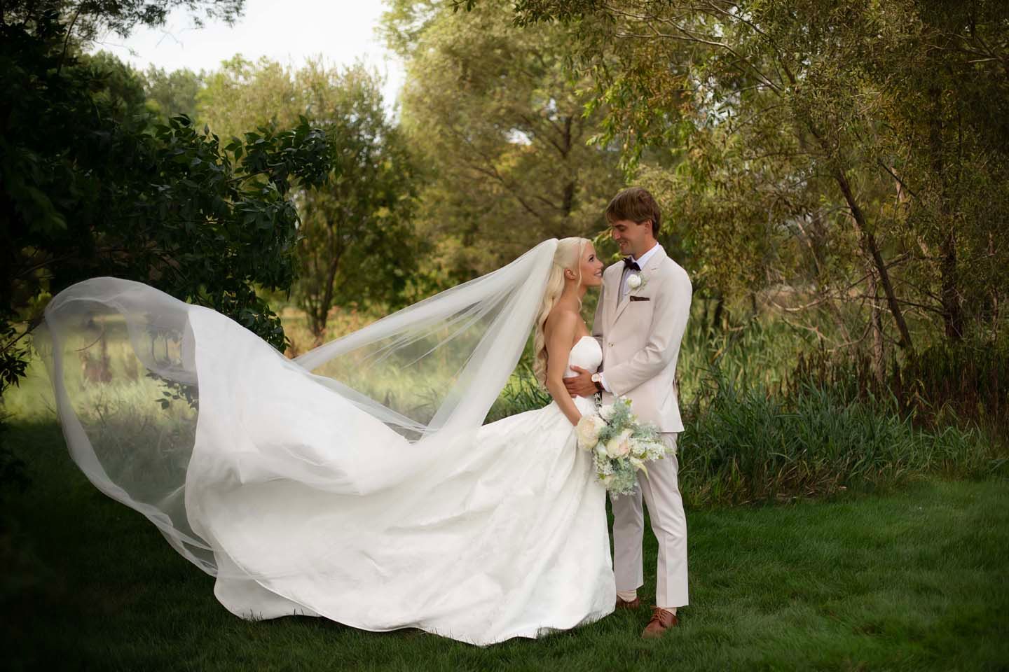 Bride and groom share a romantic kiss outdoors as her long wedding veil billows gracefully in the wind.