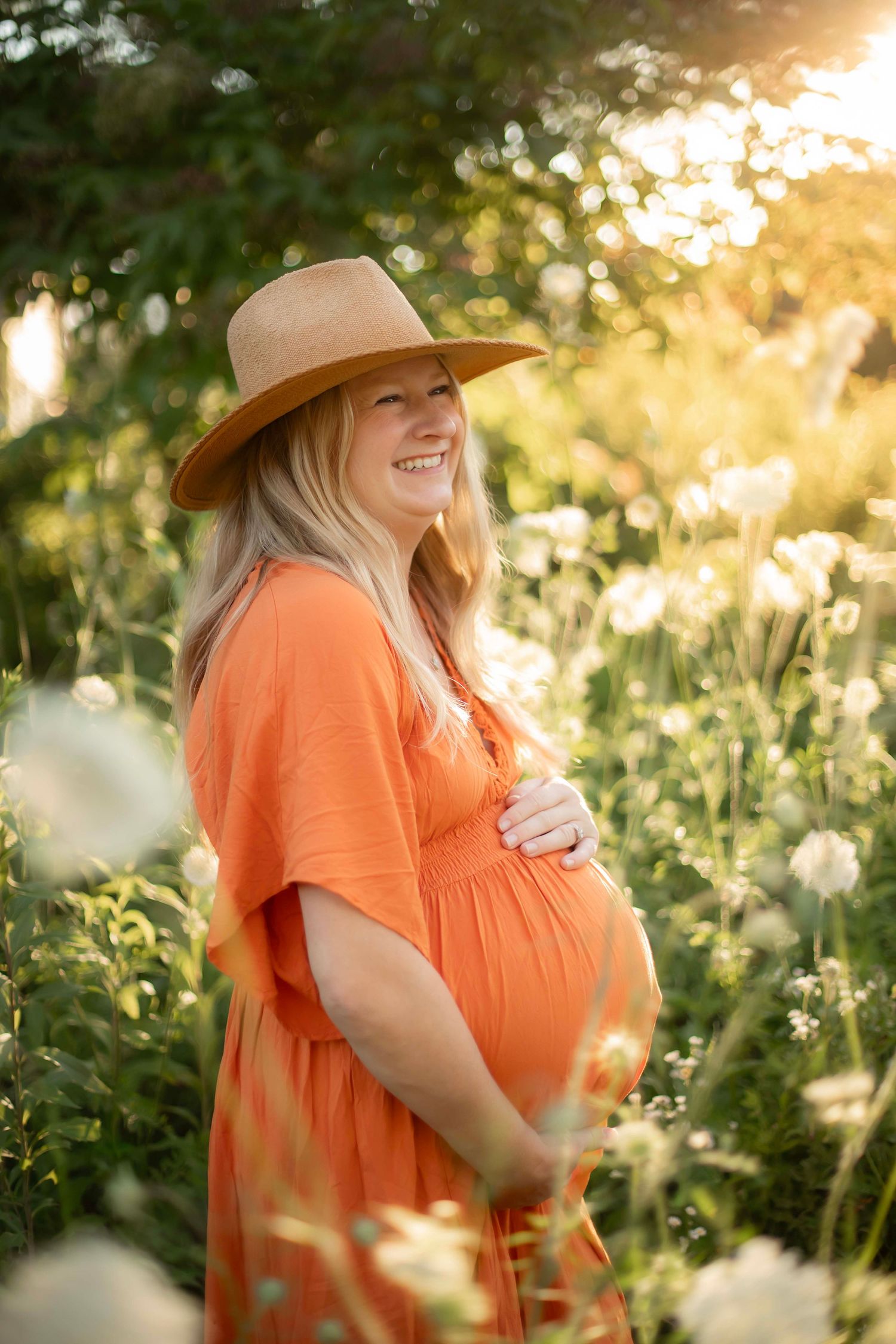Smiling pregnant woman in orange dress and tan hat stands among white wildflowers in golden sunlight.