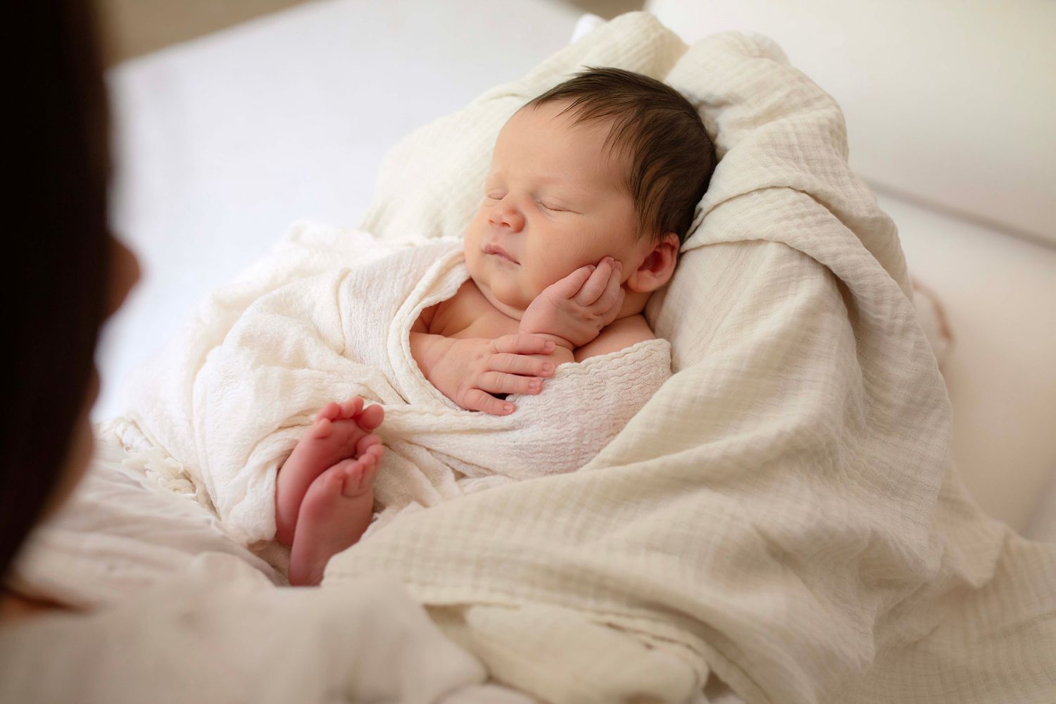 Peaceful newborn baby sleeping wrapped in soft white blanket, eyes closed with tiny hands resting on chest.