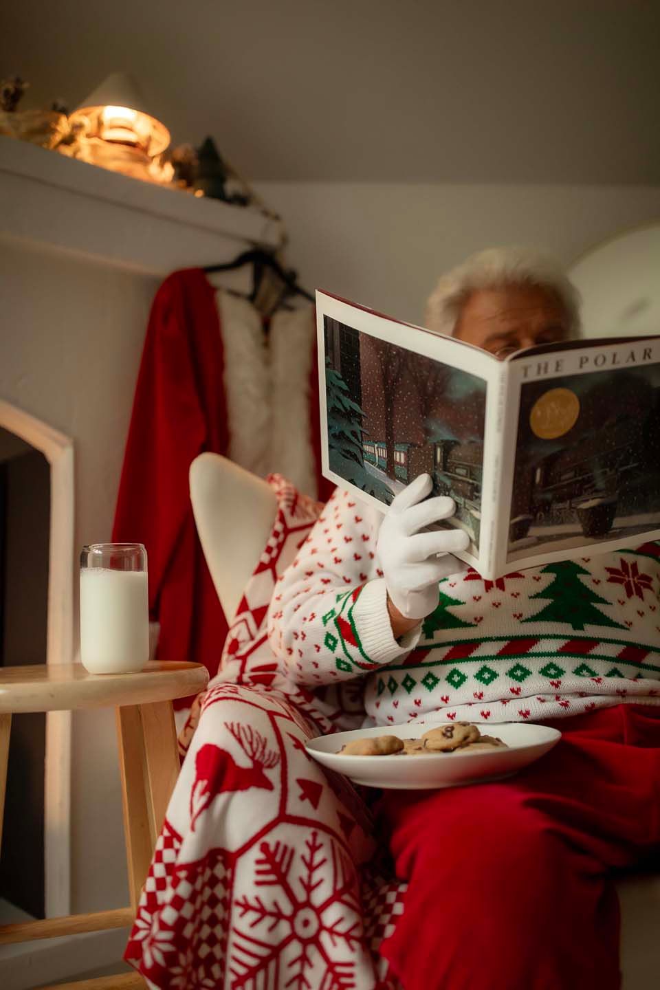 Santa Claus in festive sweater reading The Polar Express book, with milk and cookies on table nearby.