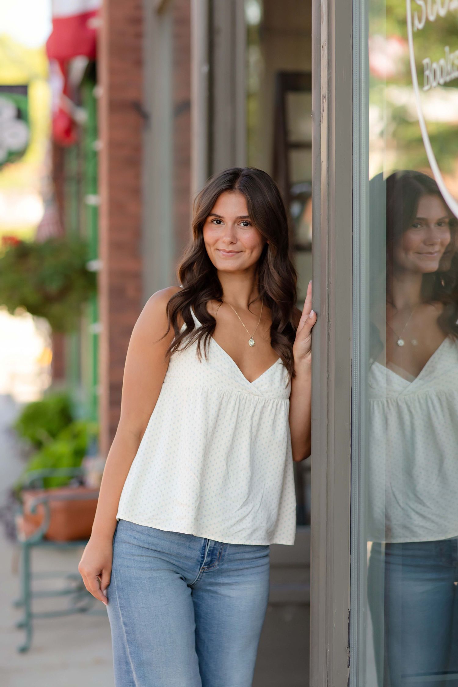 Young woman in white tank top and jeans leaning against a storefront window on a sunny day.