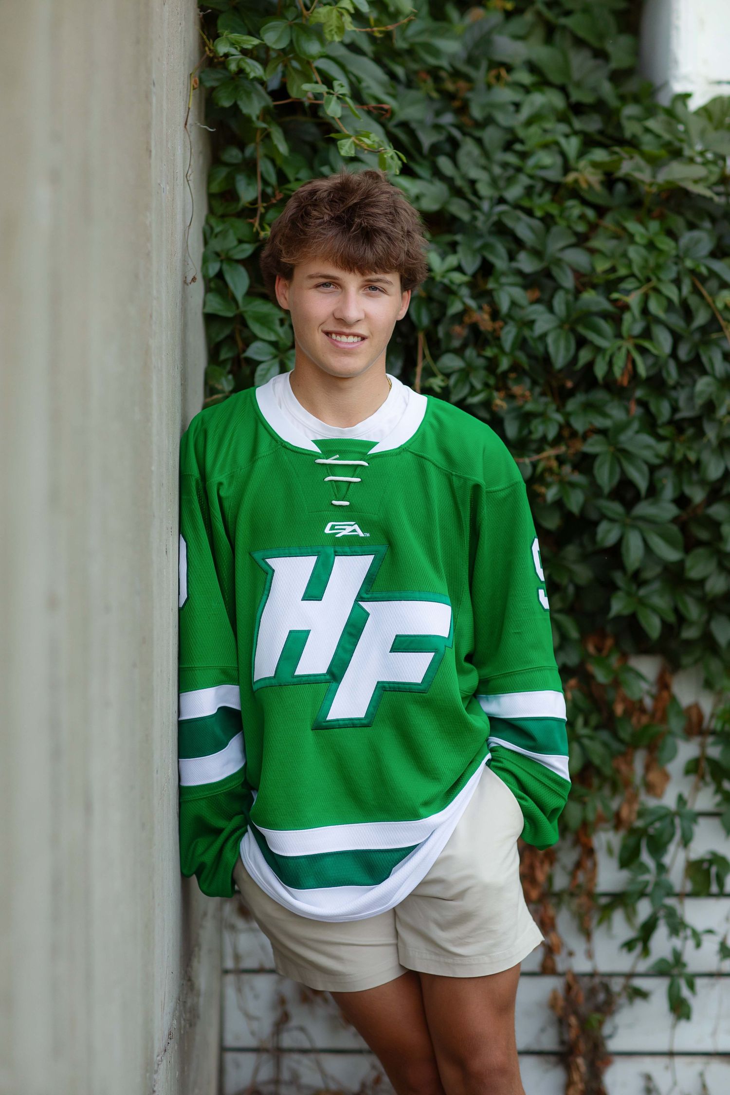 Teen boy in green HF hockey jersey smiling outdoors against ivy-covered wall.