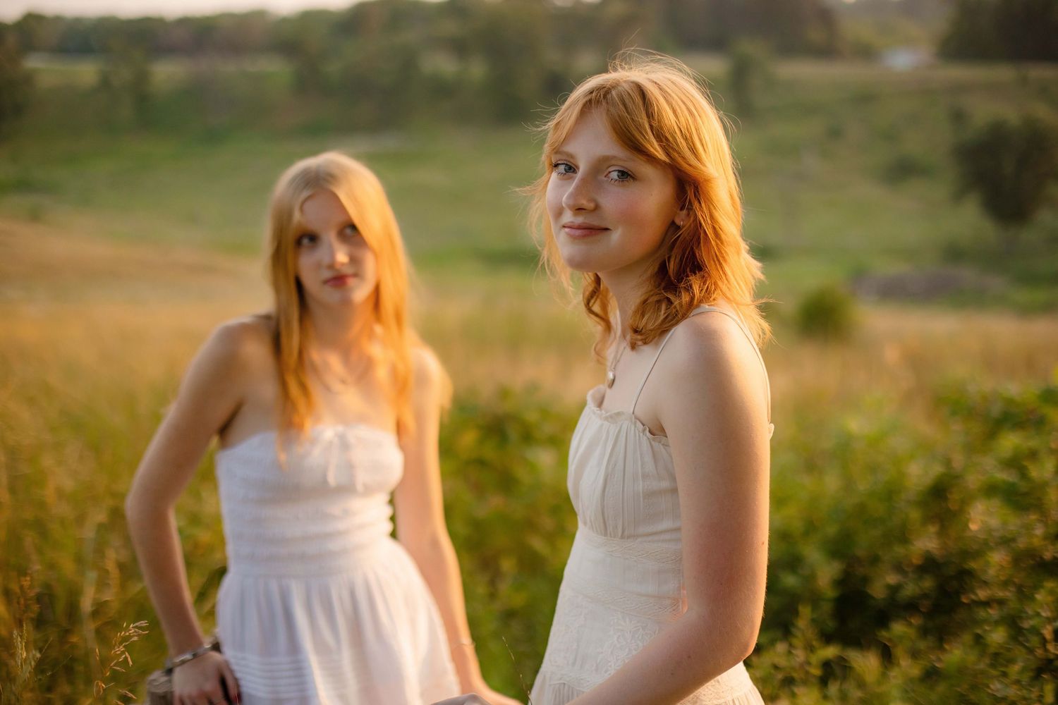 Two young women with red hair in white dresses pose in a sunlit green meadow during golden hour.