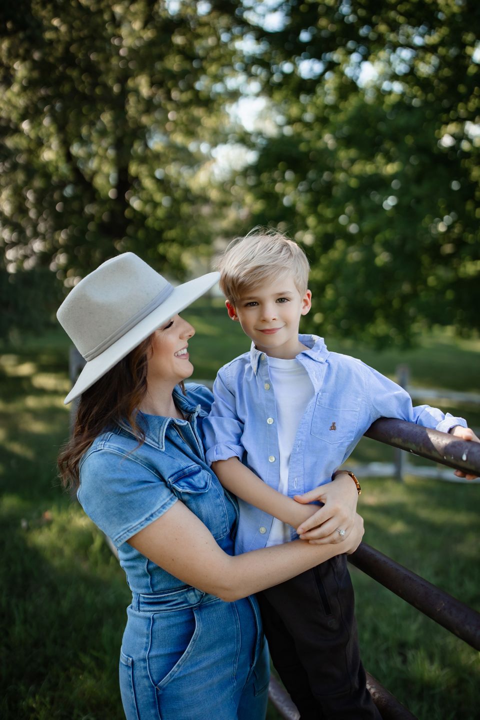 A woman in a denim outfit and white hat embraces a young blond boy in a blue shirt outdoors.