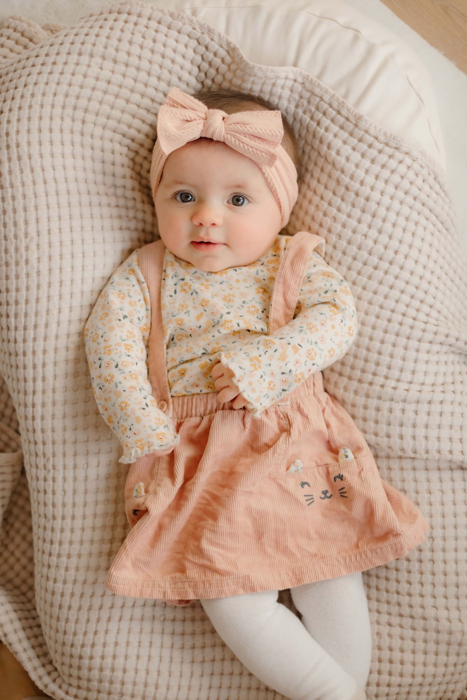 Baby in pink pinafore dress and floral top lying on textured pillow.