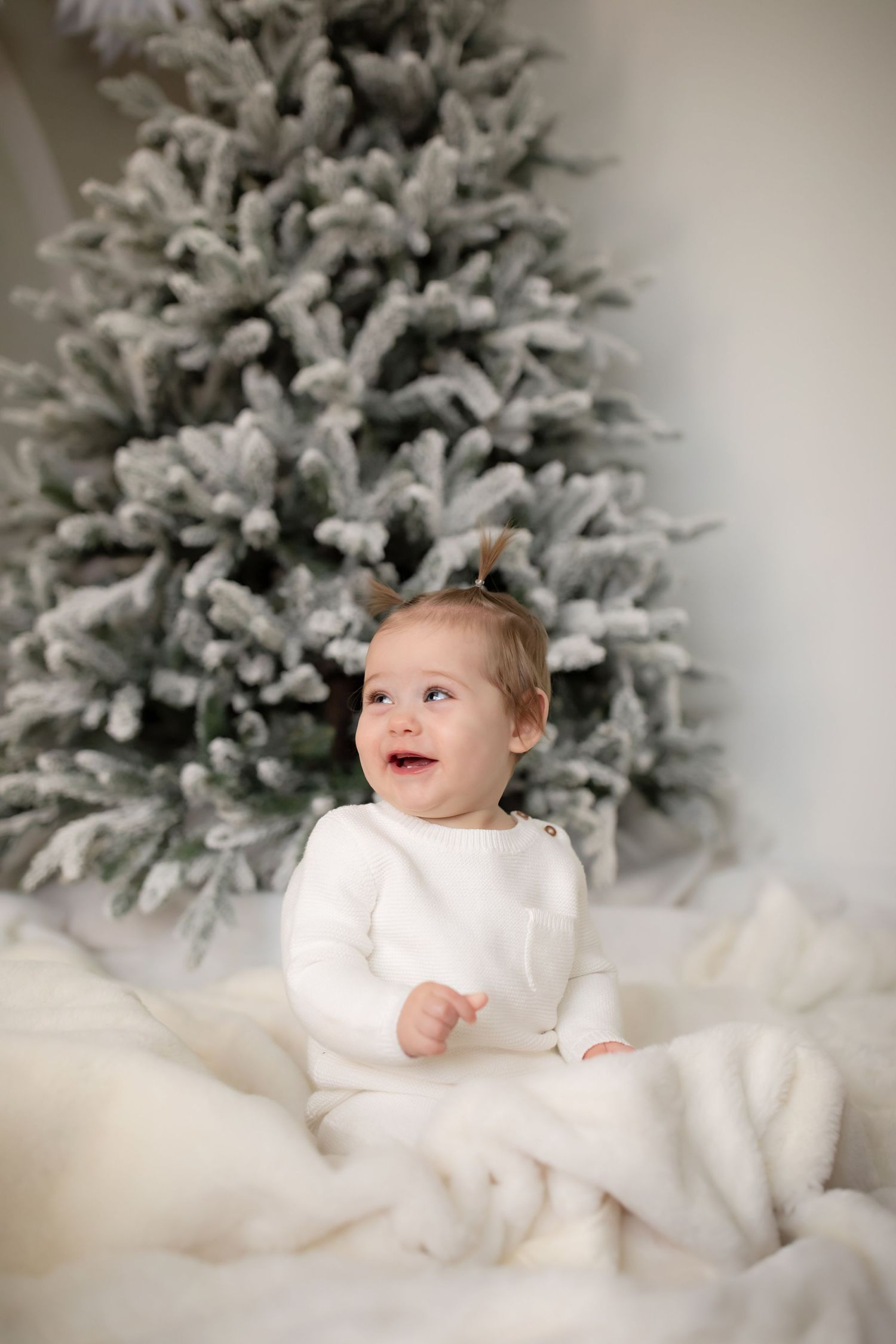 Baby in white sweater sits by snowy Christmas tree with delighted expression.