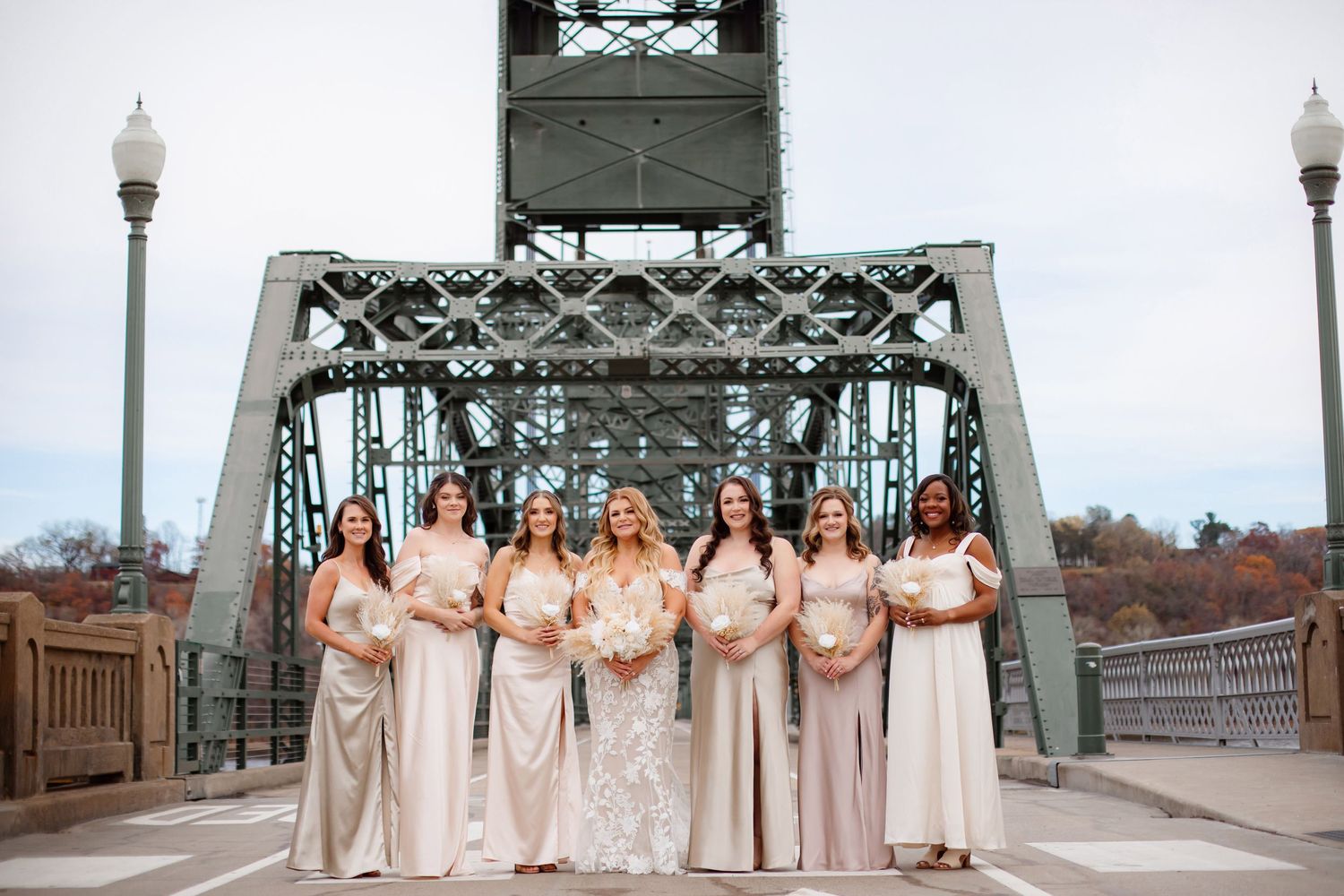 Bridesmaids in blush pink dresses pose on historic steel bridge for wedding photo.