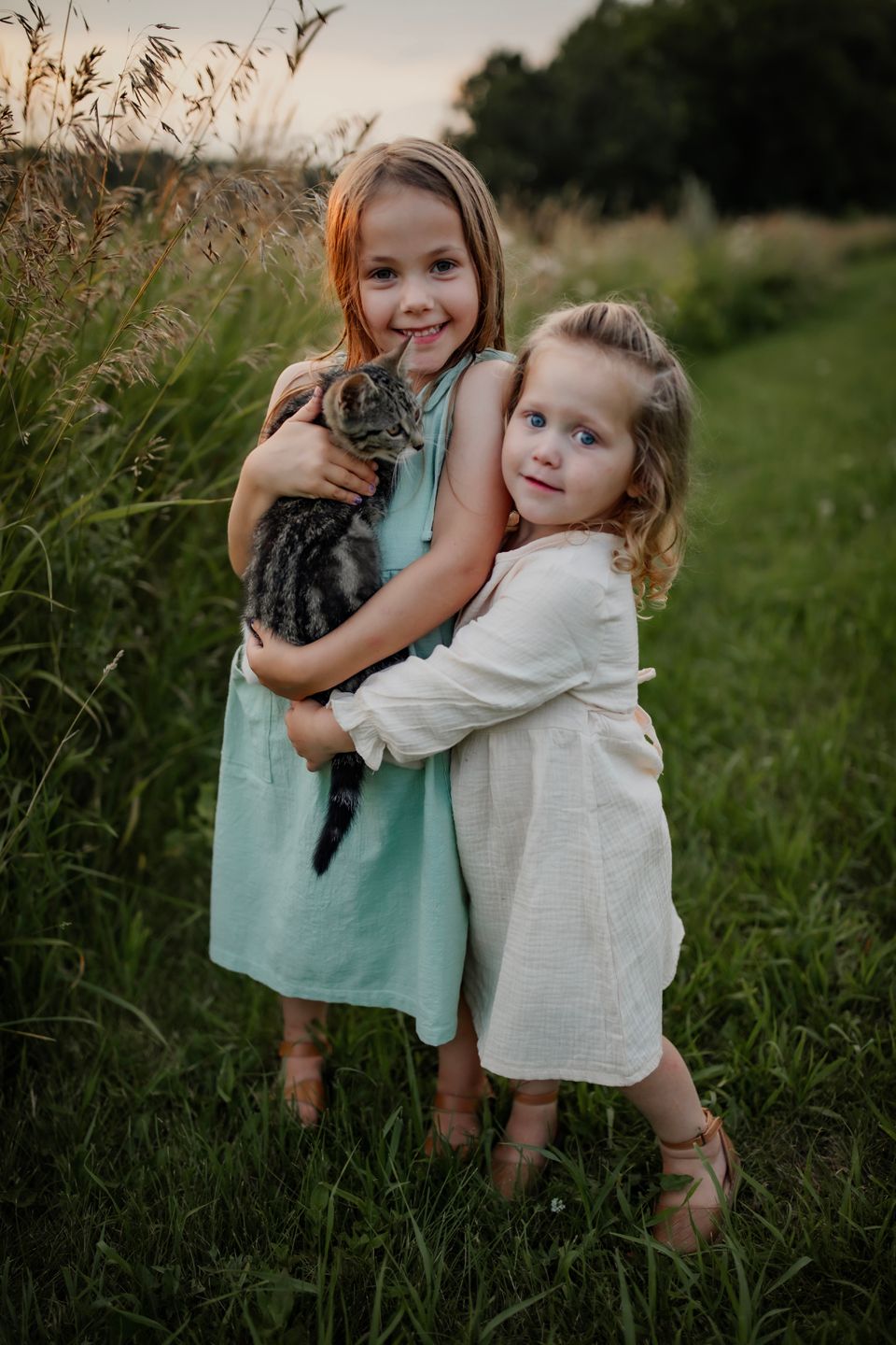 Two young friends in summer dresses hold a tabby cat in a field.