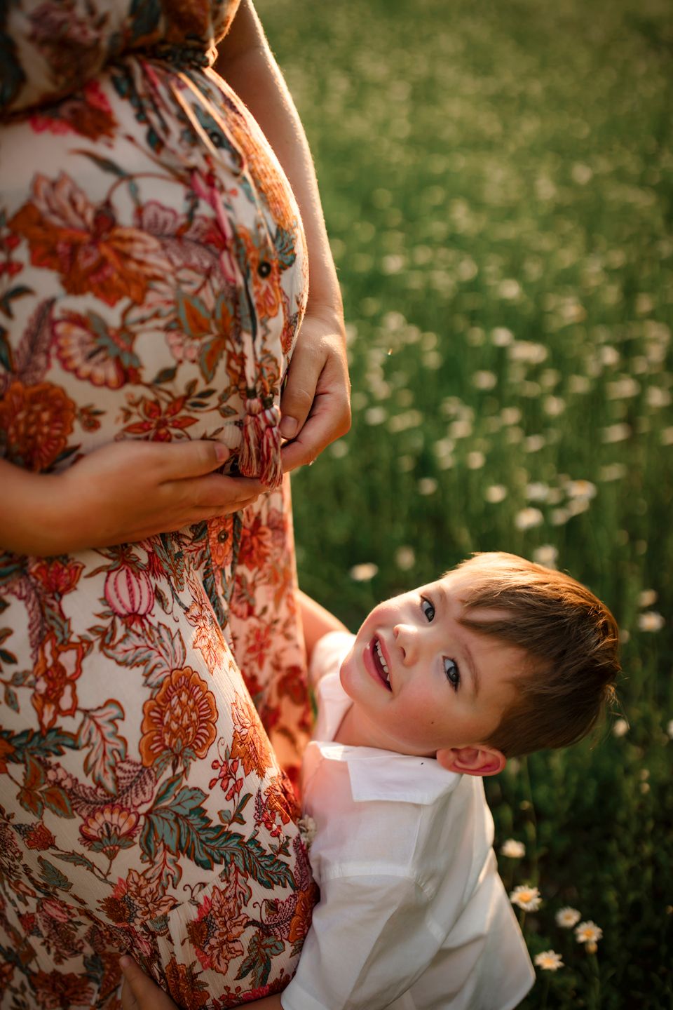 Low angle view of young child hugging someone wearing a floral orange dress in a field of daisies at sunset.