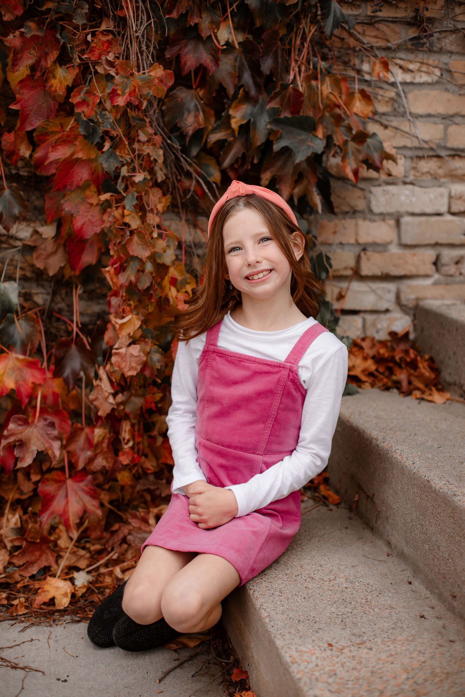 Portrait in pink jumper dress against brick wall with fall ivy backdrop.