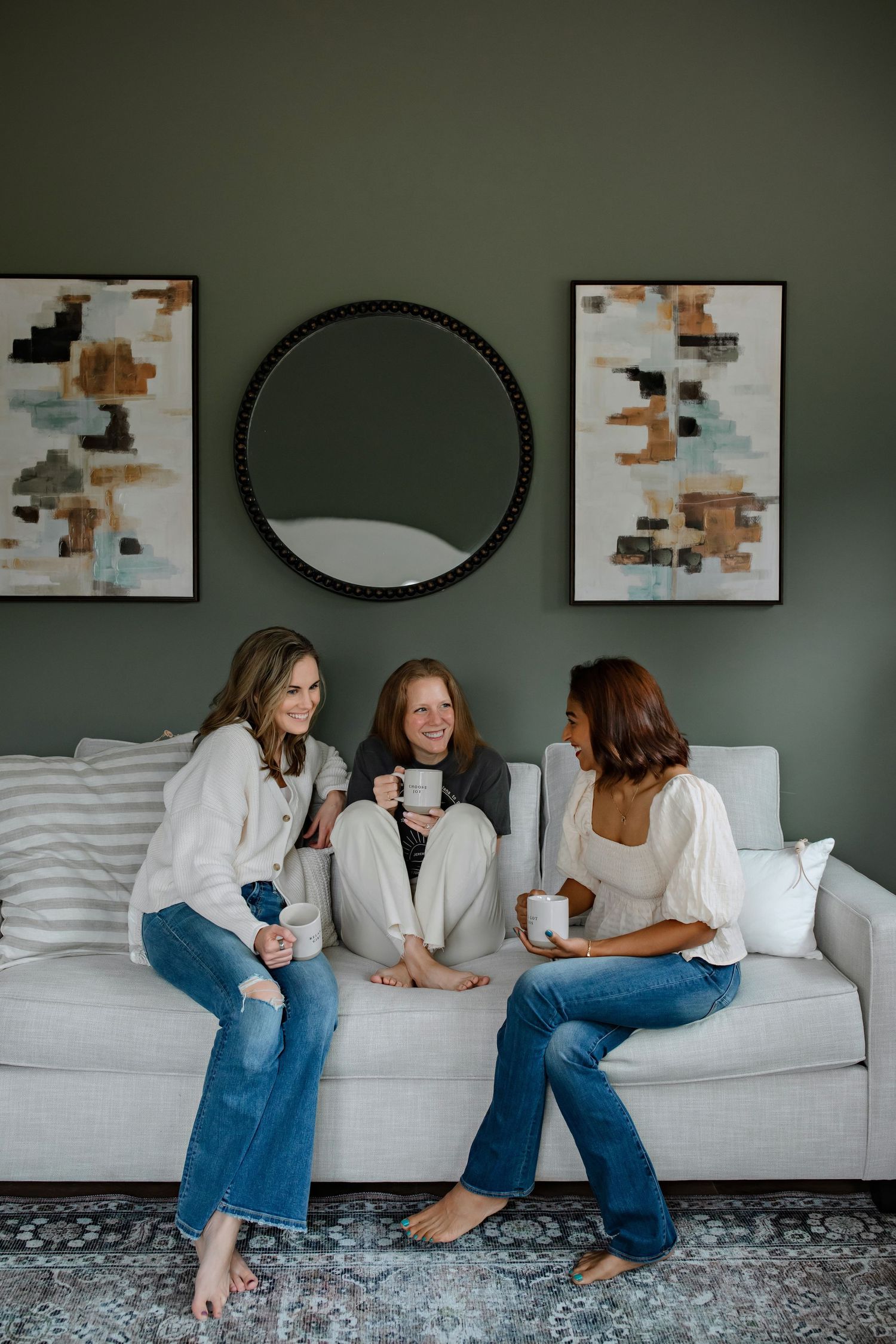 Three friends chatting on a gray couch with abstract art and round mirror on sage green wall.