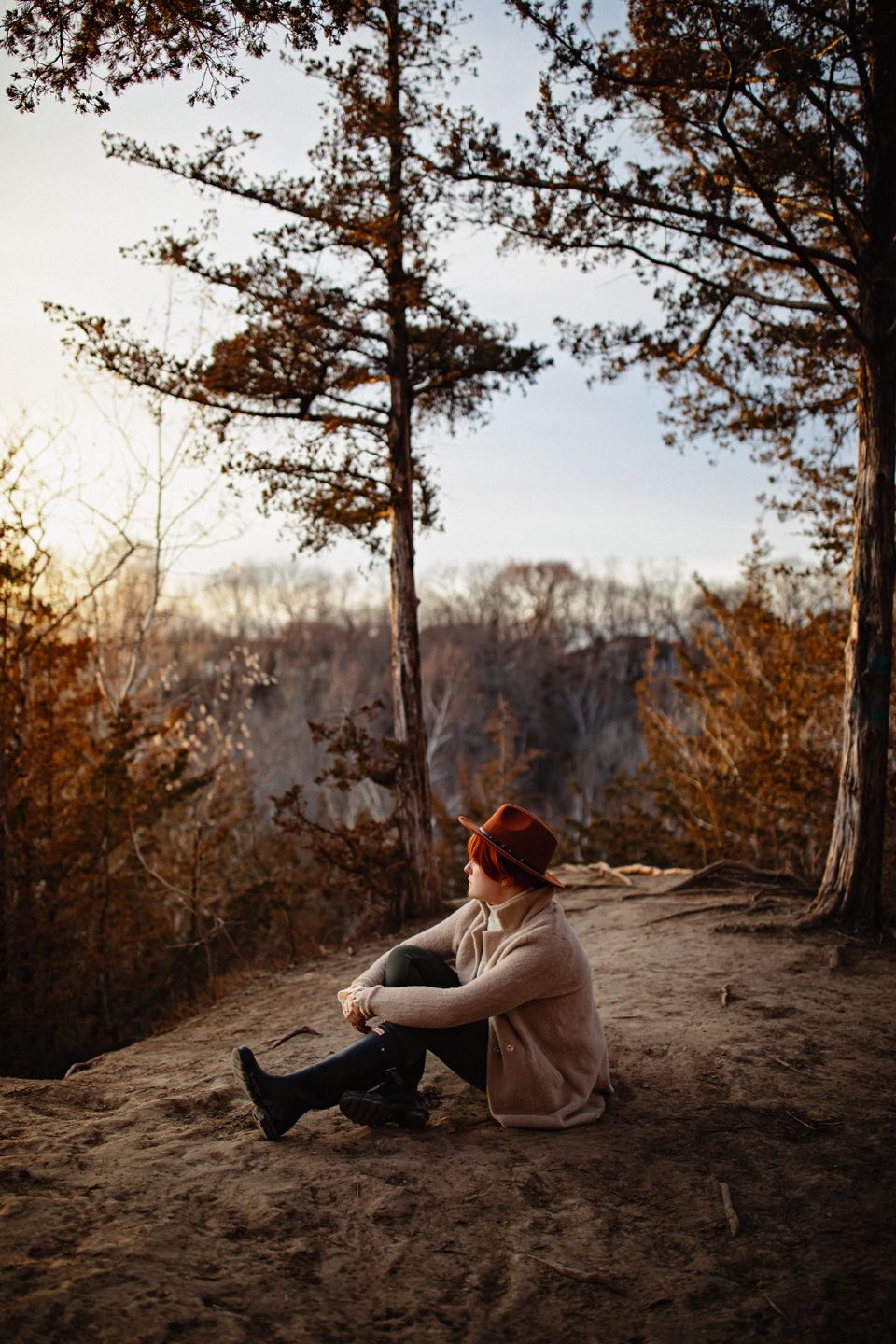 A solitary figure sits on a clifftop overlooking a forest during sunset.