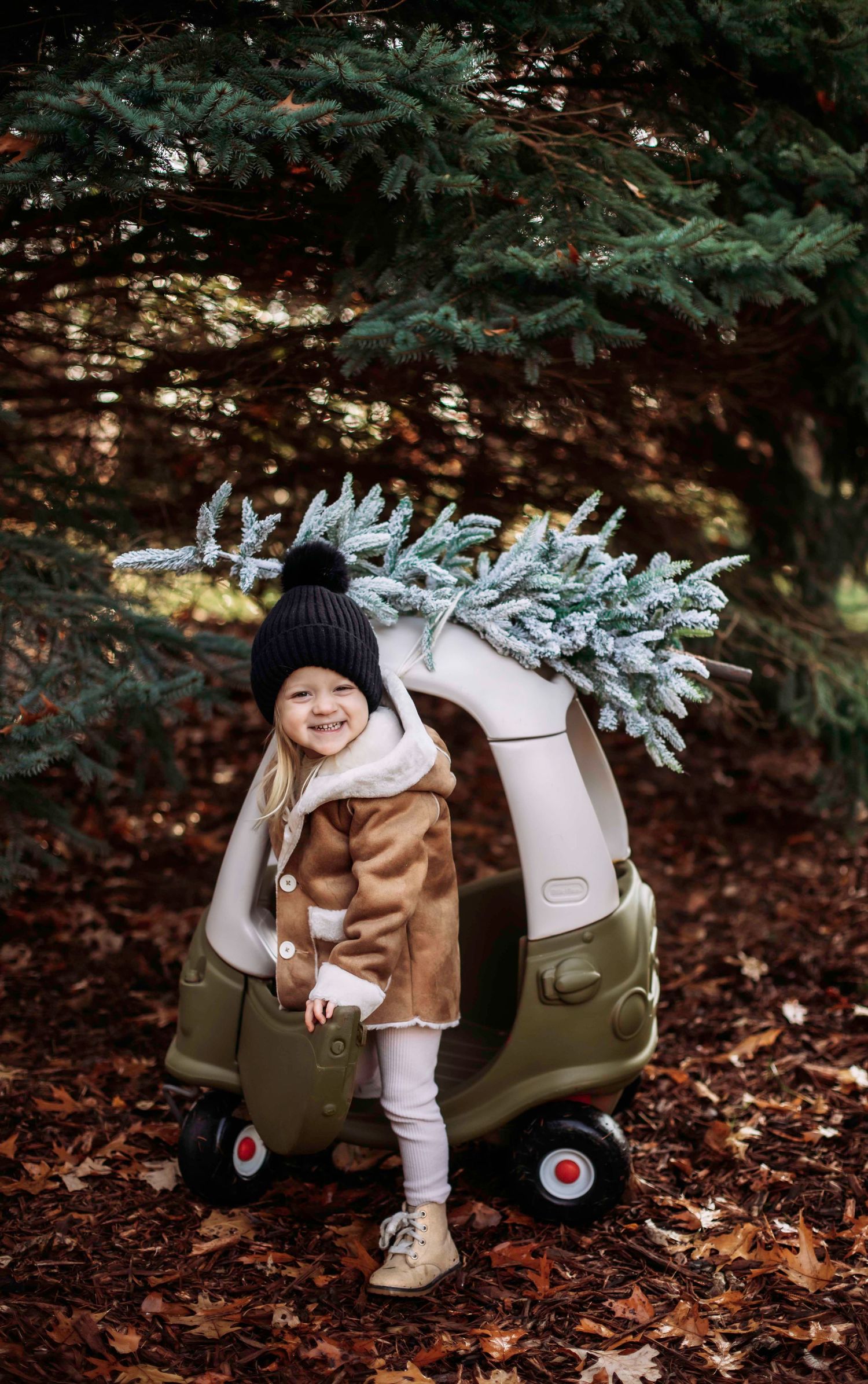 Decorated toy cozy coupe with festive greenery and child in winter attire in autumn forest.
