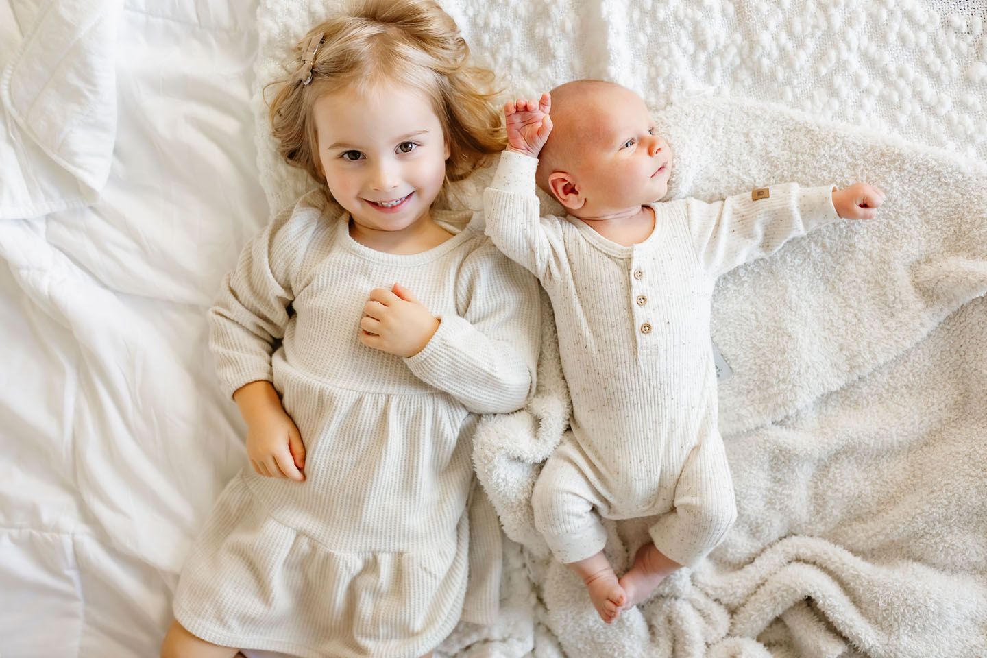 Two children in matching cream outfits lying together on white bedding.