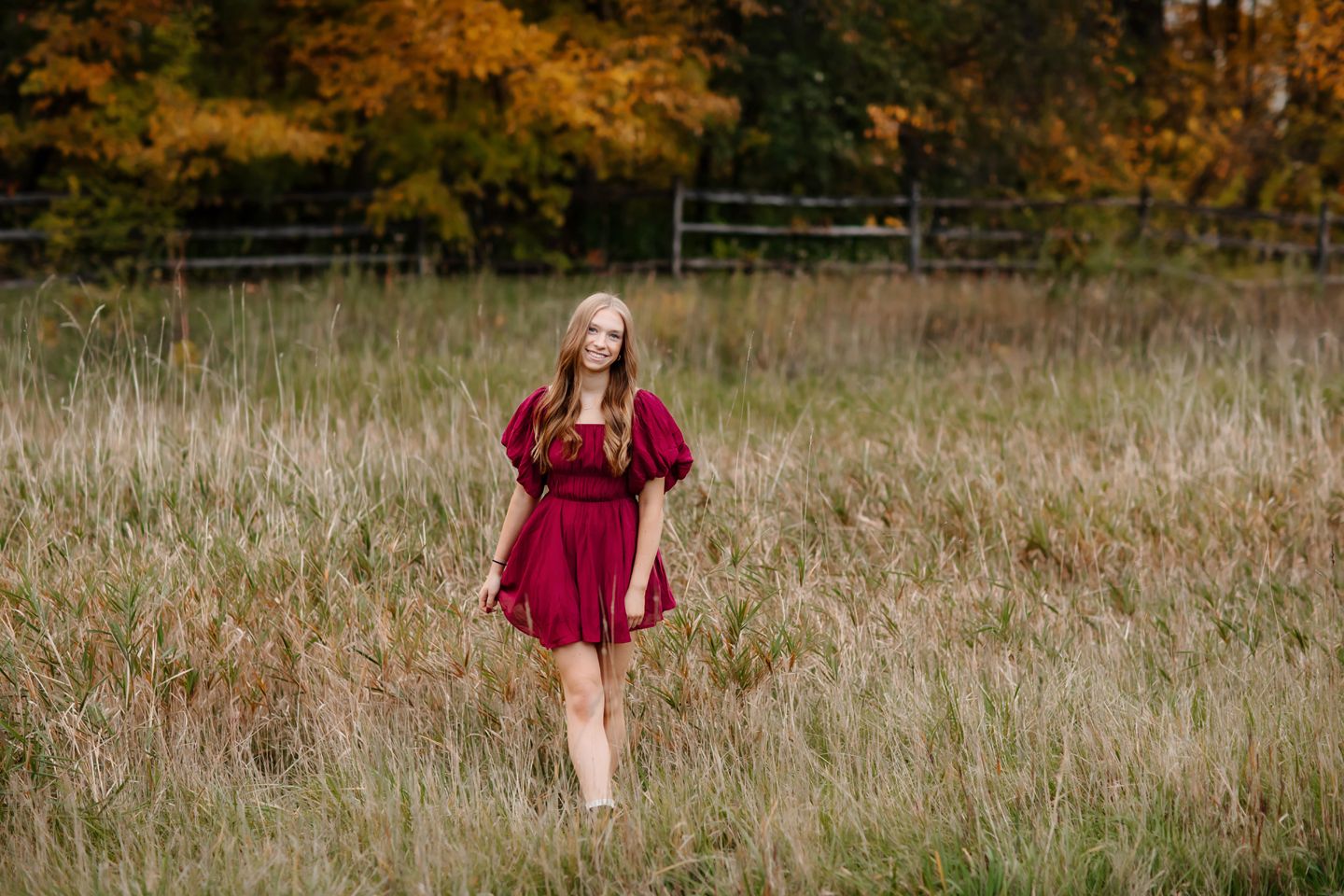 Flowing burgundy dress contrasts beautifully against tall autumn grasses and wooden fencing.