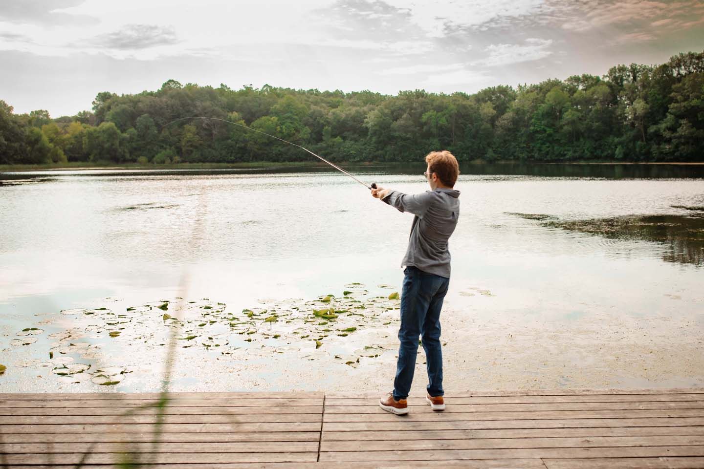 Someone fishing from wooden dock at sunset over peaceful lake surrounded by trees.