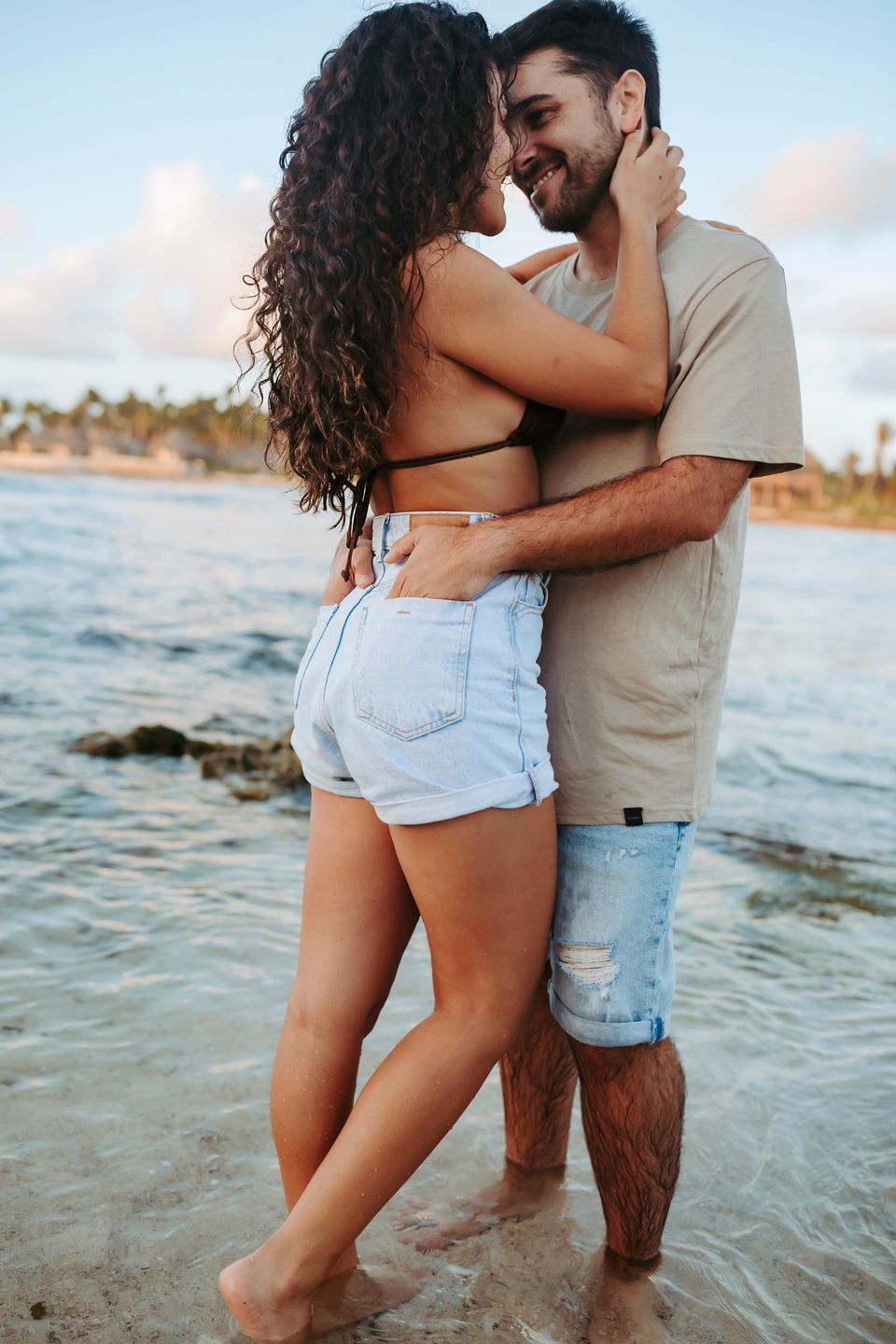 A tender moment between a couple standing in shallow water at a rocky beach during sunset.
