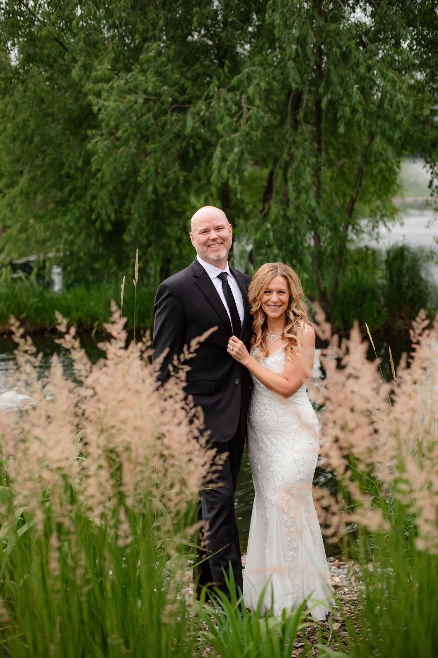 Romantic wedding couple poses in a garden setting with flowing ornamental grasses and willow trees.