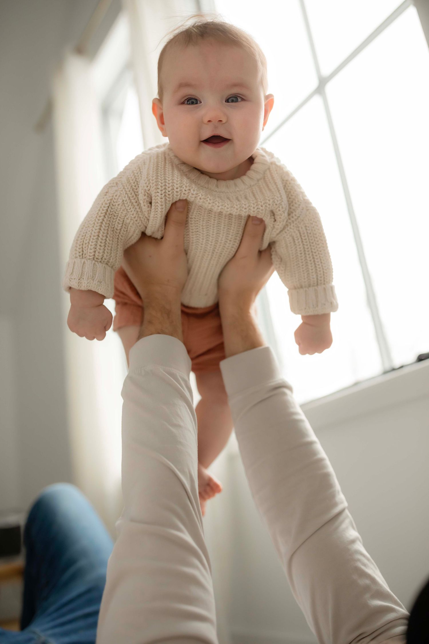 A baby wearing a cream-colored sweater is lifted up and held in the air against a bright window background.