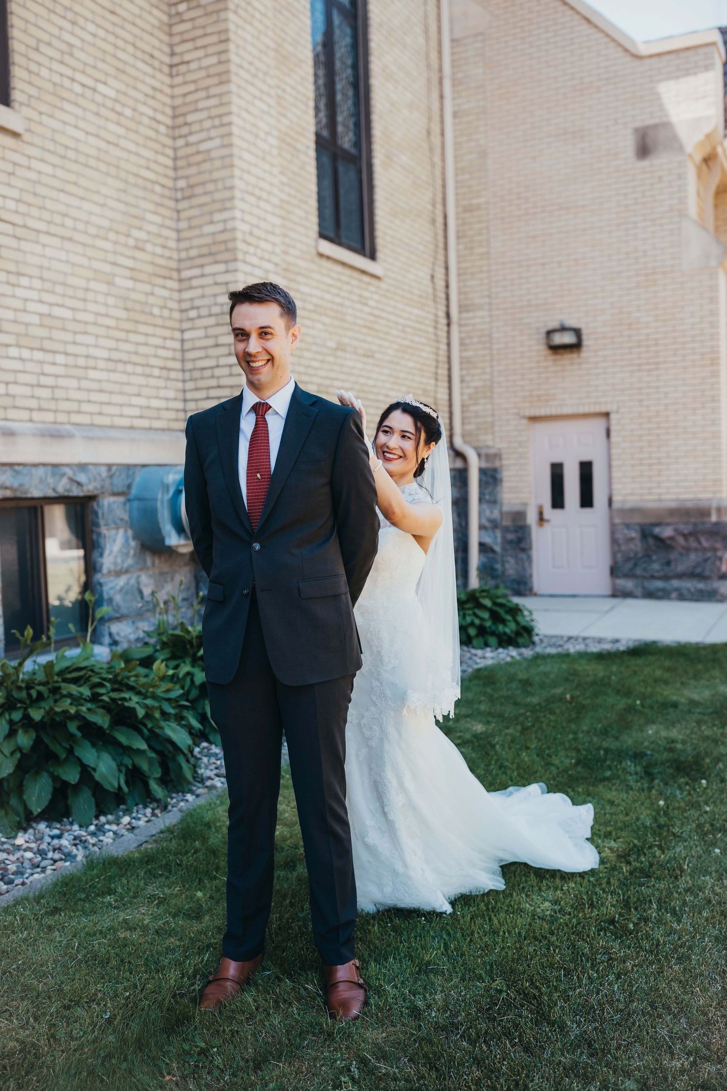 Playful wedding photo outside brick church with couple doing first look pose.