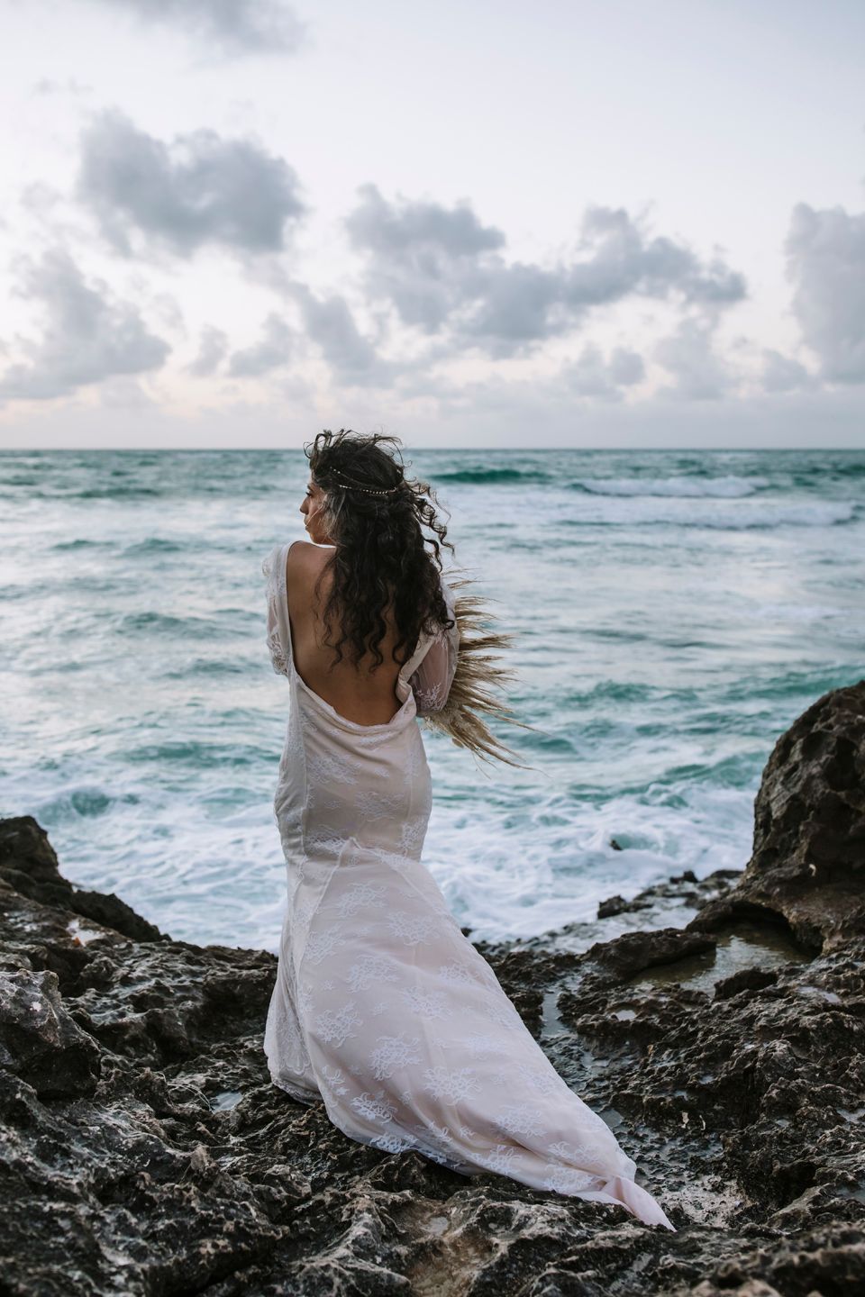 A bride in backless white dress stands on rocky shoreline facing the dramatic ocean waves.