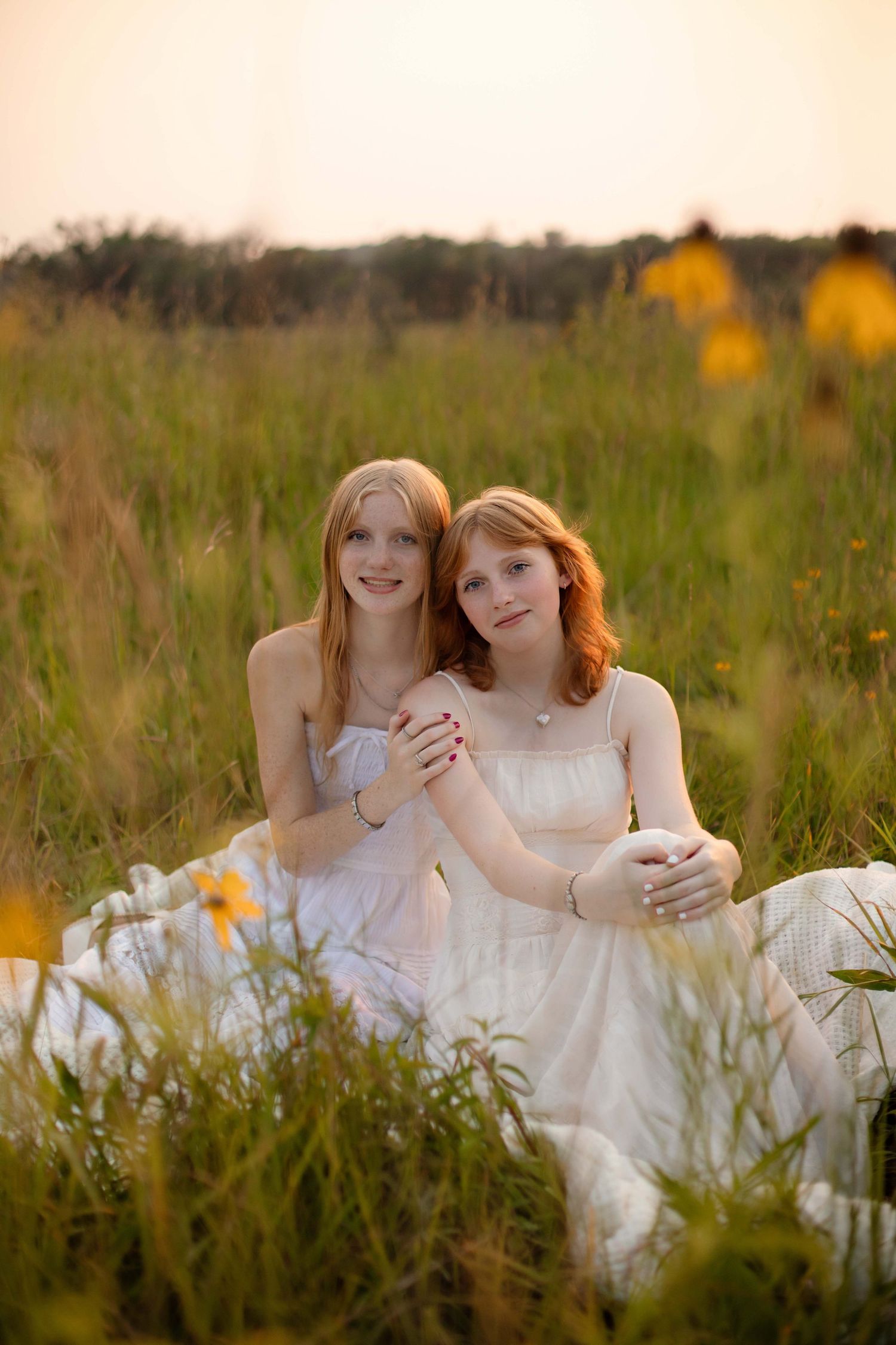 Two redhead girls in white dresses sit together in a golden meadow at sunset, smiling warmly.