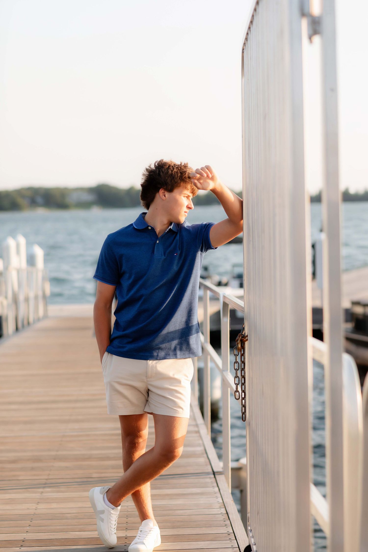 Young man in blue polo and khaki shorts leaning on marina dock railing with waterfront view.