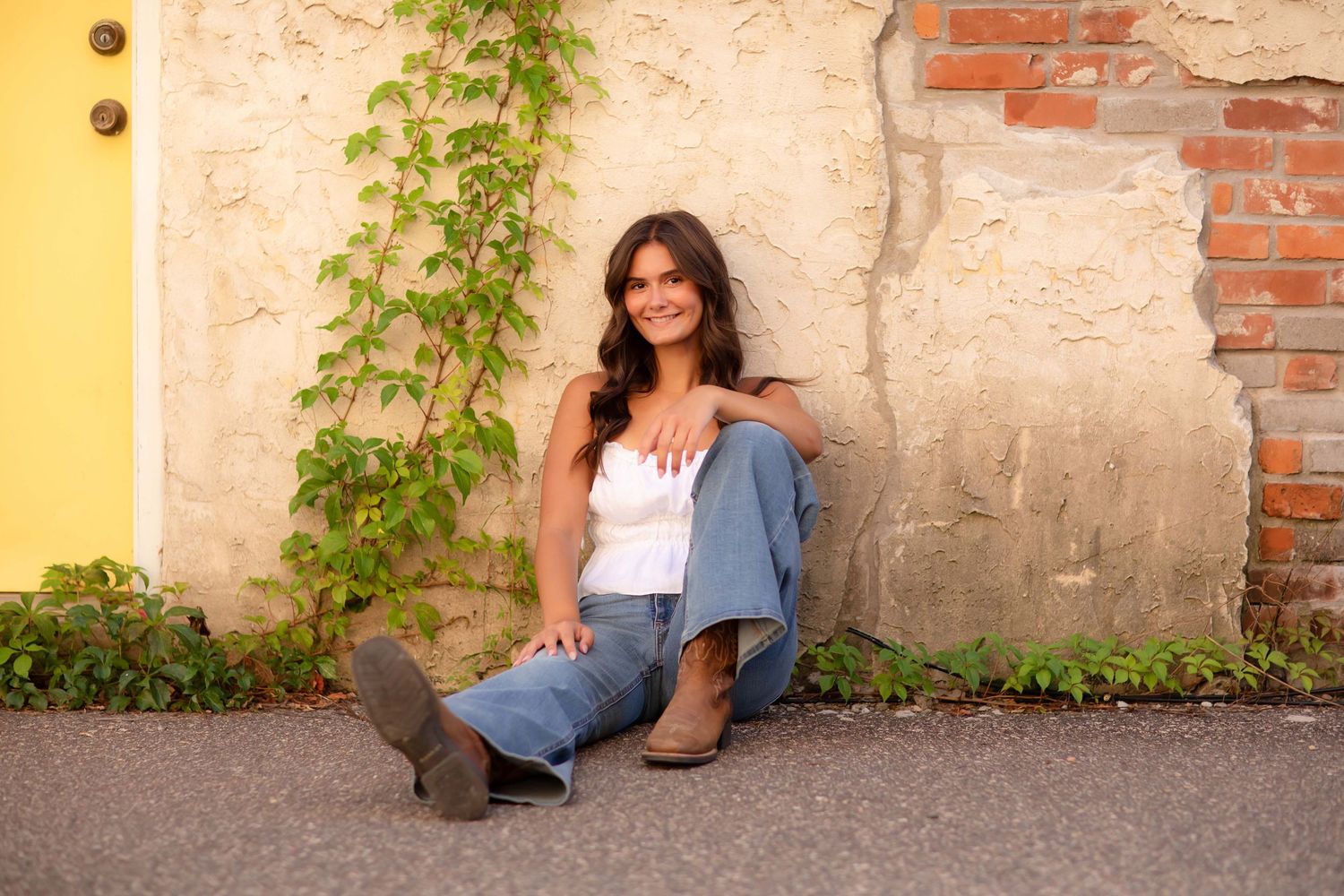 Woman in white top and jeans sitting against rustic wall with ivy, smiling outdoors.
