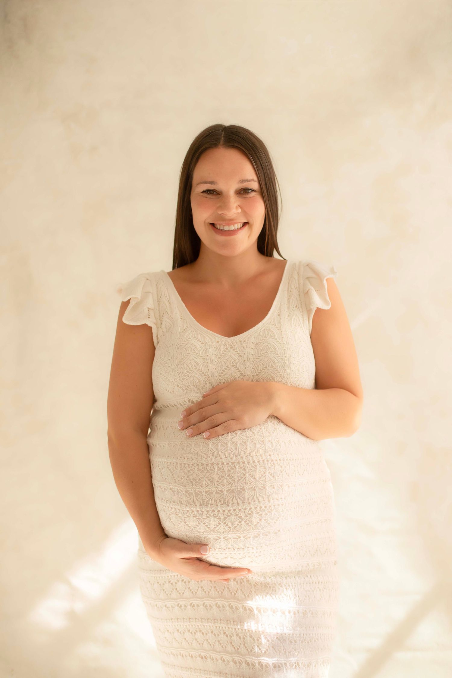 Smiling pregnant woman in white lace dress gently holding her baby bump indoors.