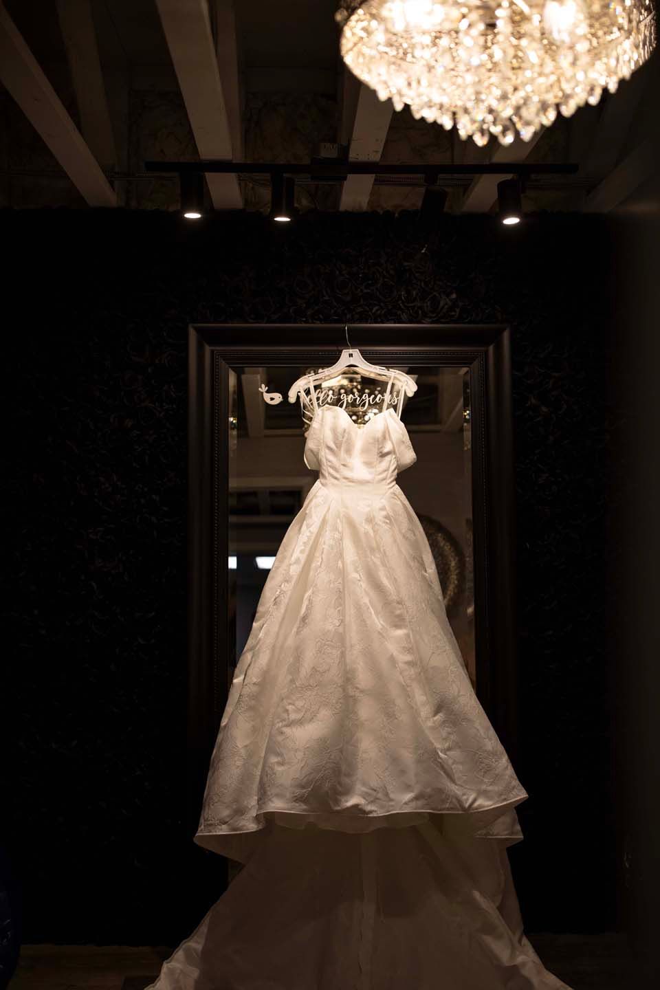 Elegant white off-shoulder wedding gown displayed on a hanger beneath a crystal chandelier in a dimly lit bridal boutique.