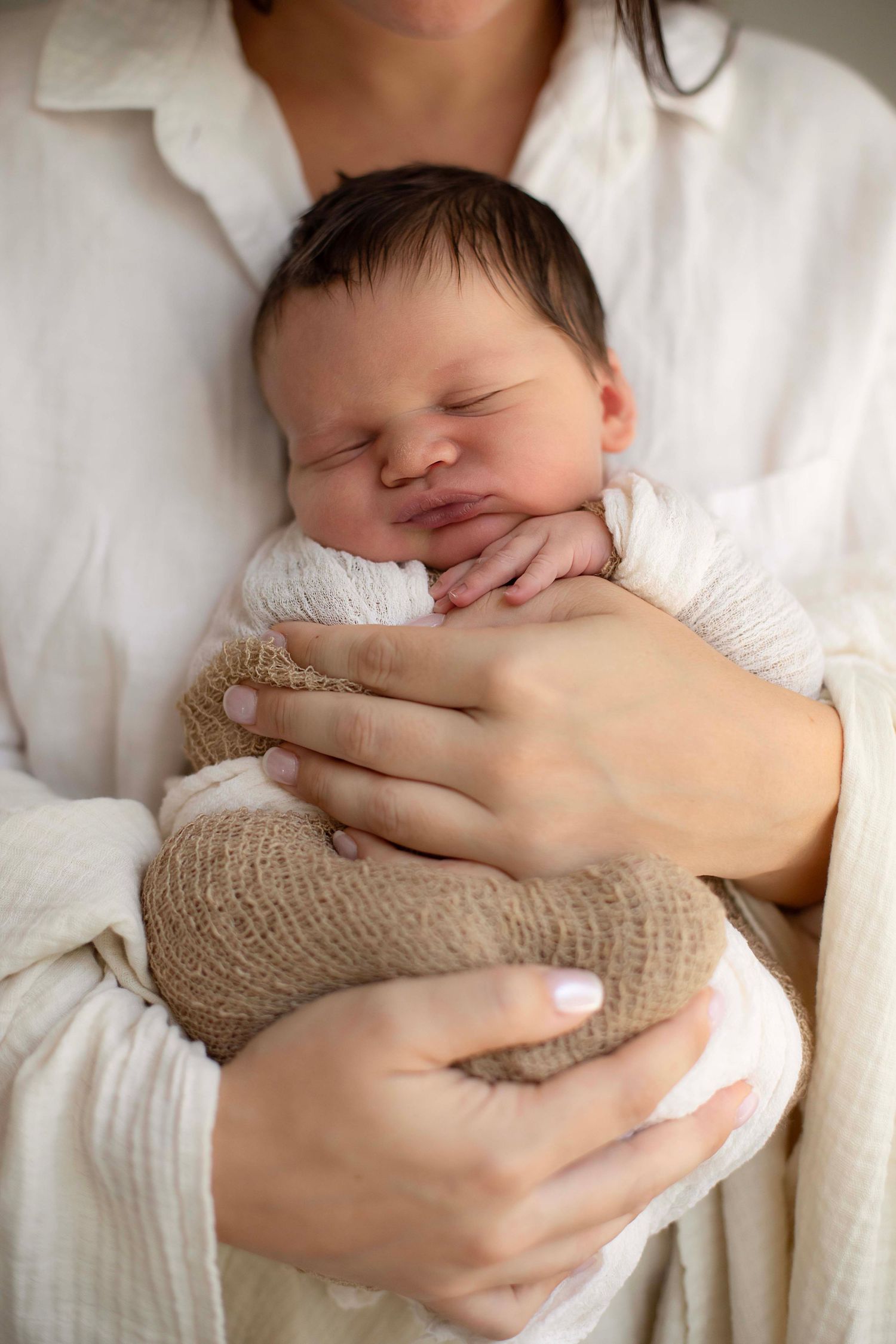 A mother gently cradles her sleeping newborn baby wrapped in white and burlap swaddle cloths.