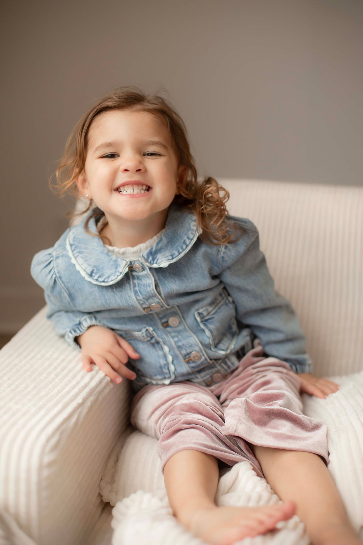 A child in a denim jacket and pink shorts reclines playfully on a white couch in a series of candid poses.