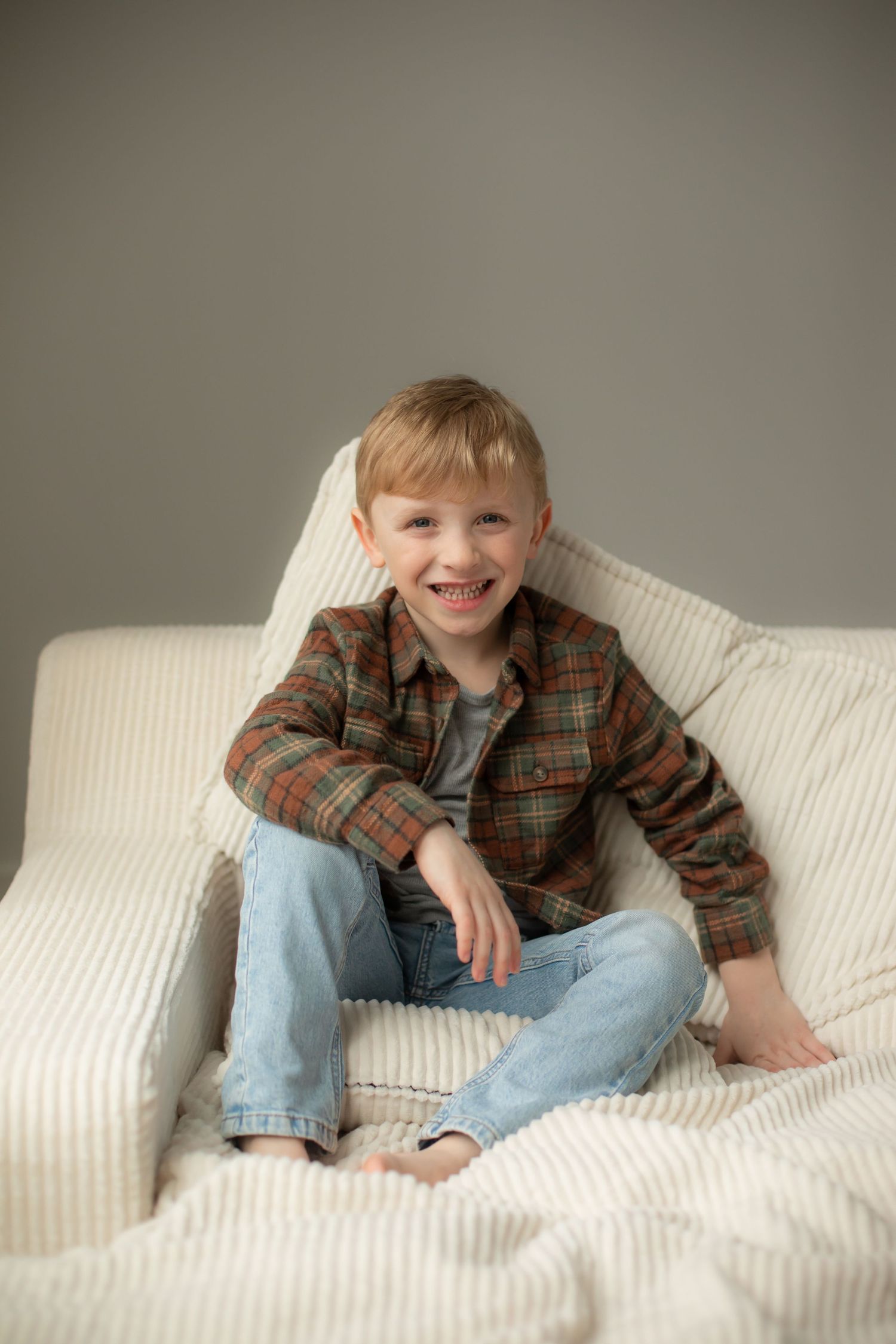 Child wearing plaid shirt and jeans relaxes on white couch in casual indoor photoshoot series.