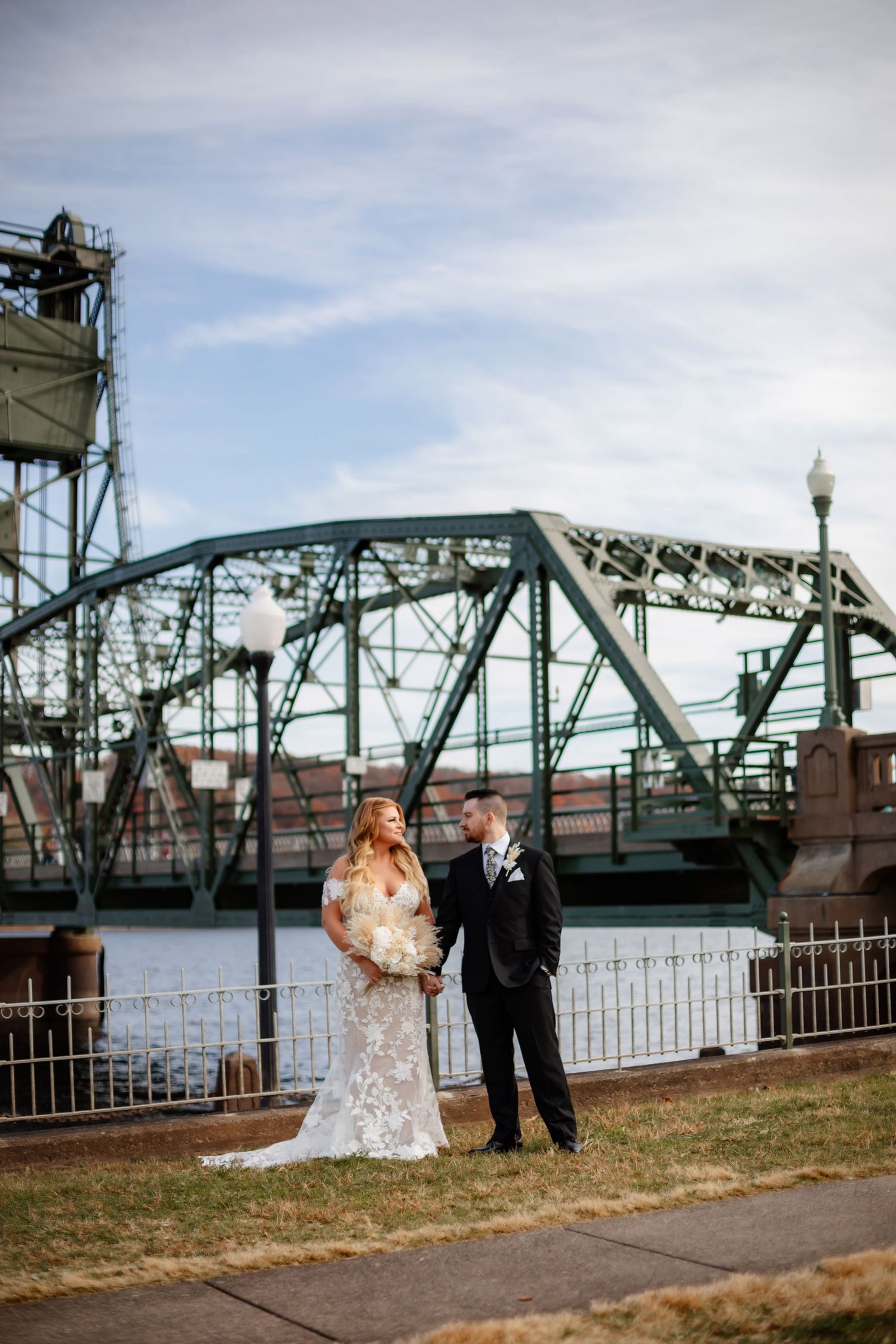 A wedding couple poses in front of a large steel bridge structure on a cloudy day.