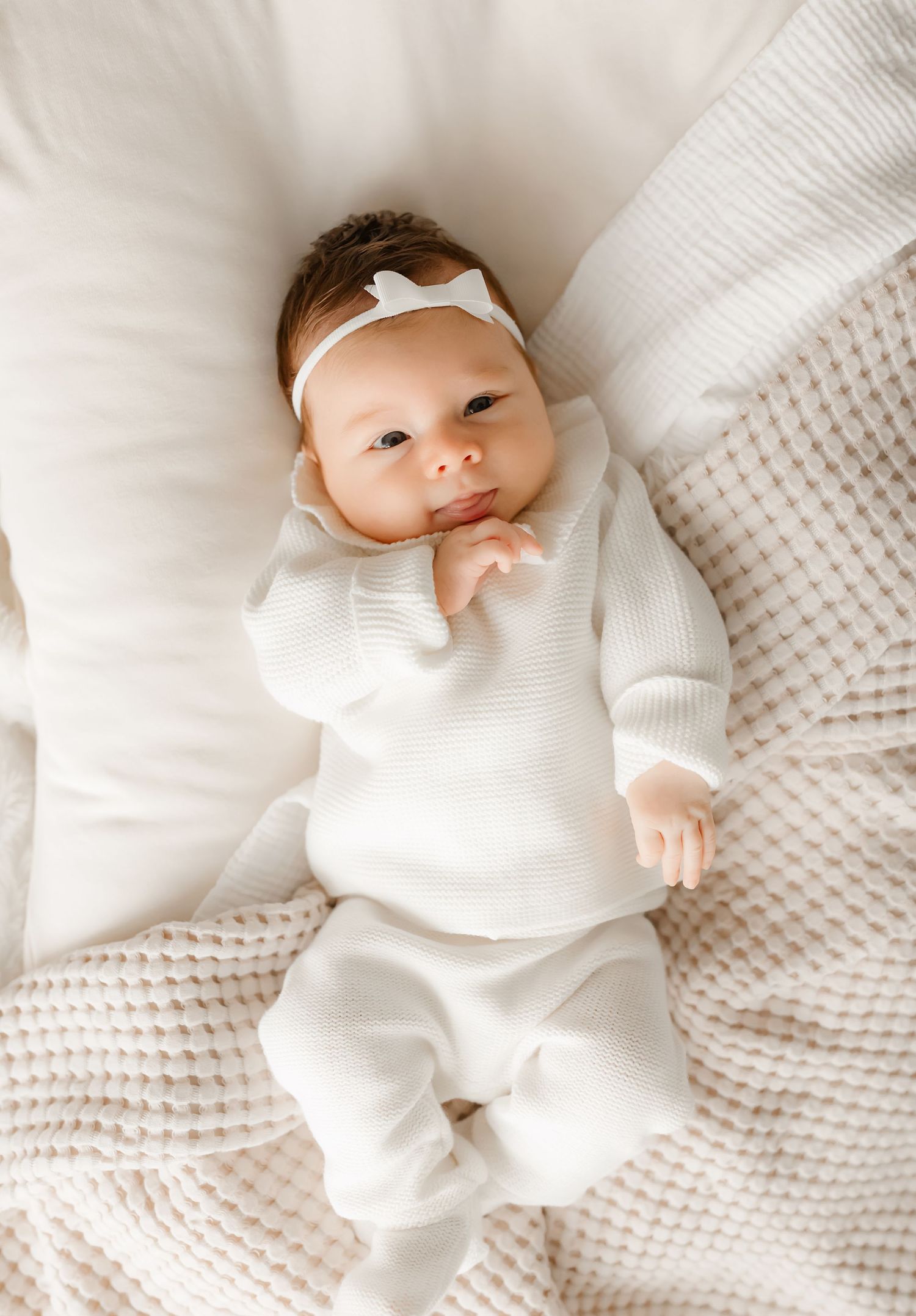 Baby in white knit outfit and headband lying on textured blanket.