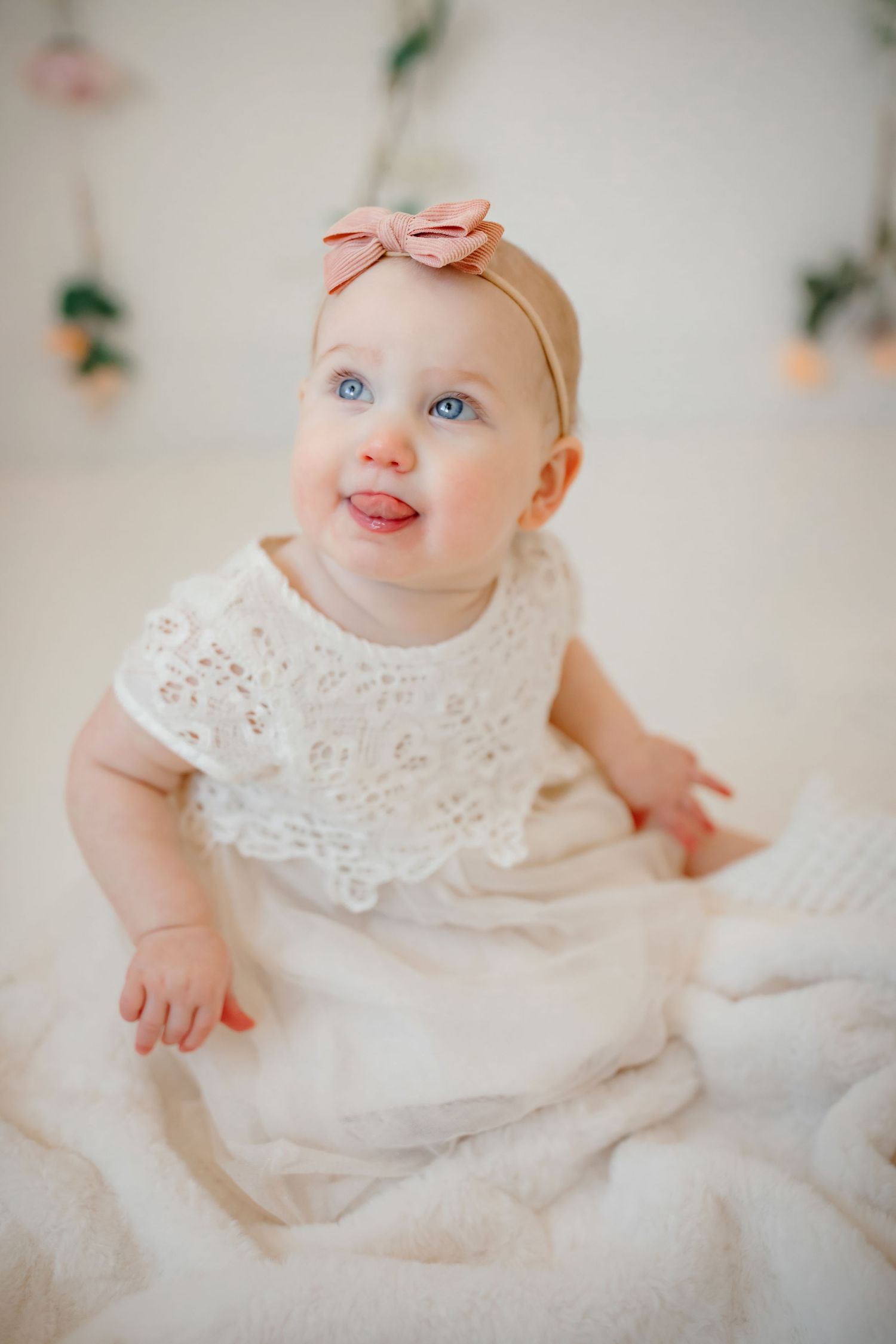 Baby in white lace dress with peach bow sitting on white fabric.