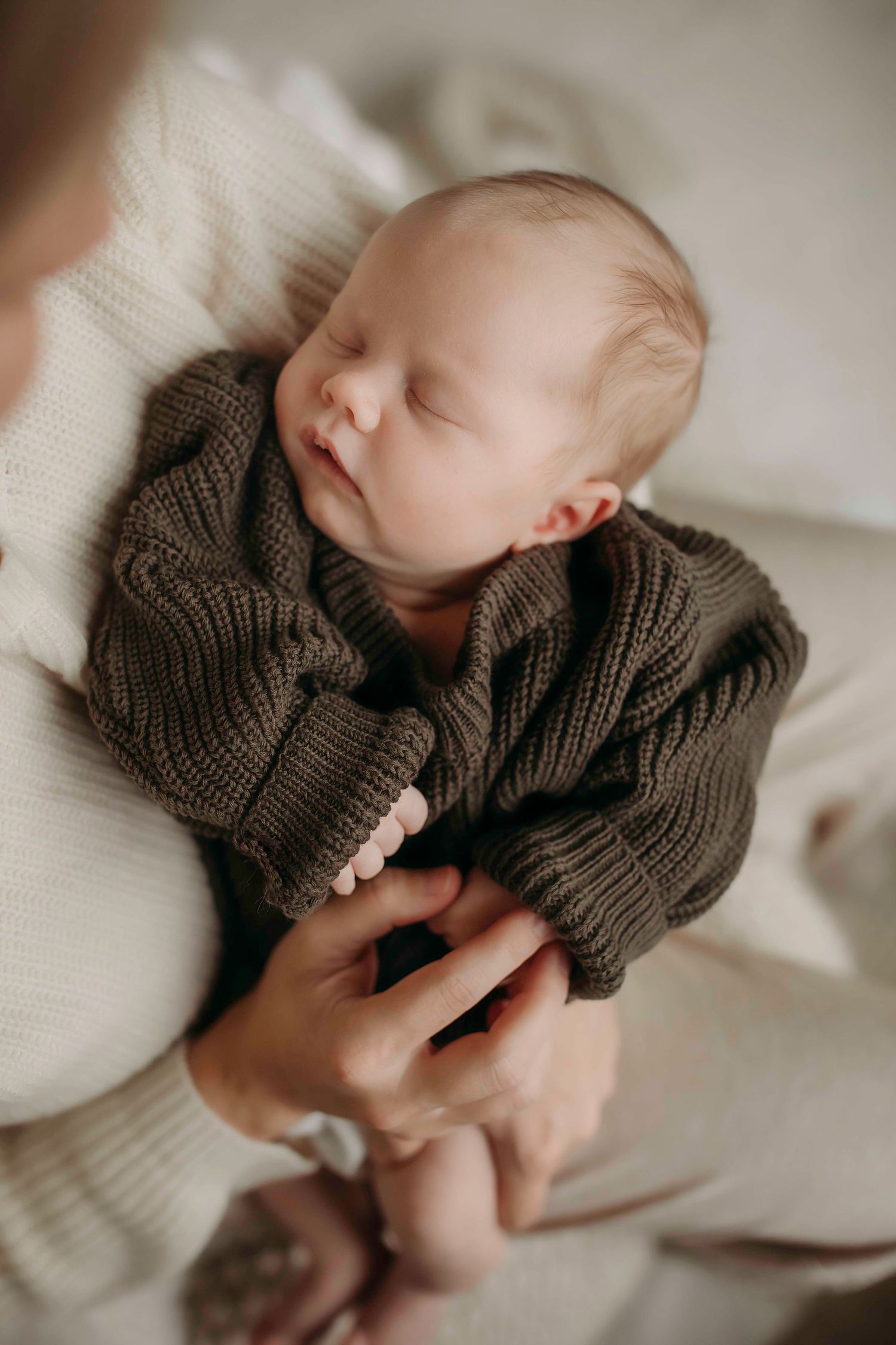 Sleeping baby wearing brown knit sweater held in caregiver's arms.