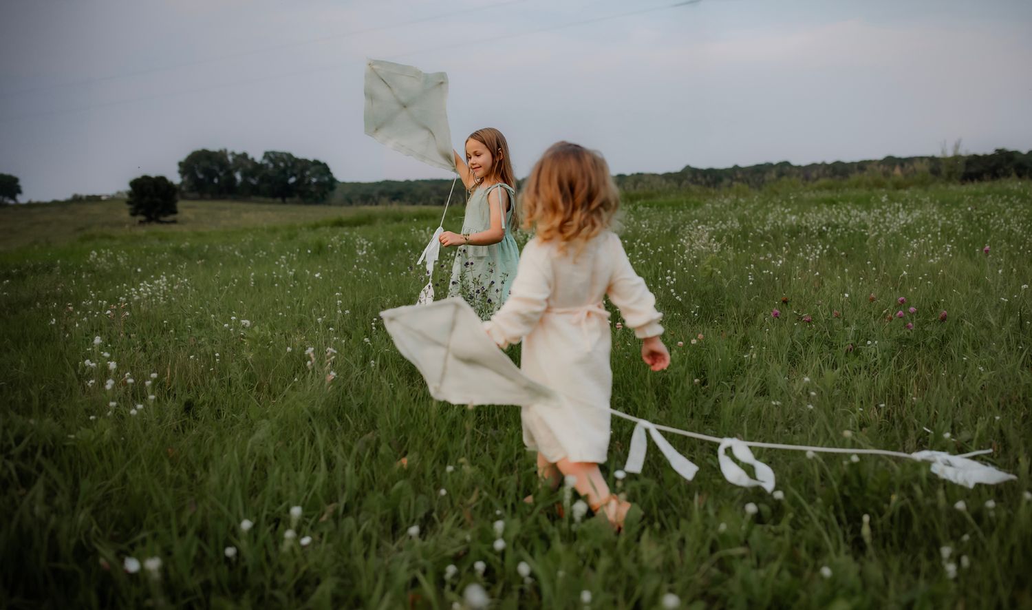 Two children running through a wildflower meadow with handmade kites on a cloudy day.