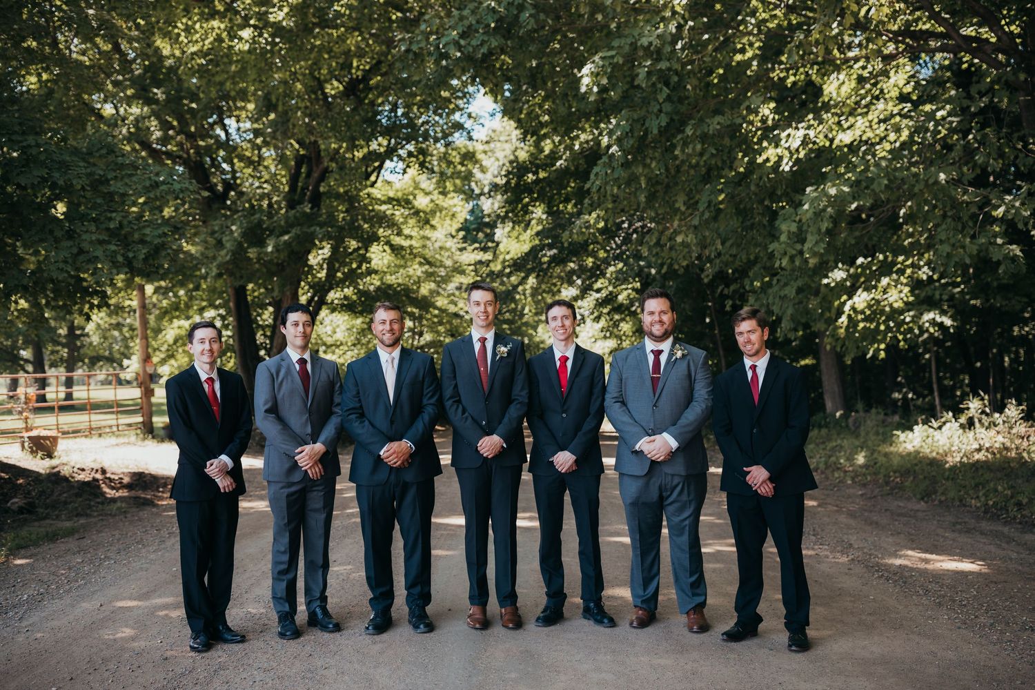 Wedding party in formal suits with red ties posed outdoors on a tree-lined path.