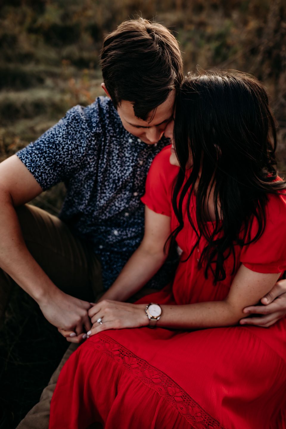 Romantic couple in navy floral shirt and red dress holding hands during outdoor golden hour photoshoot.