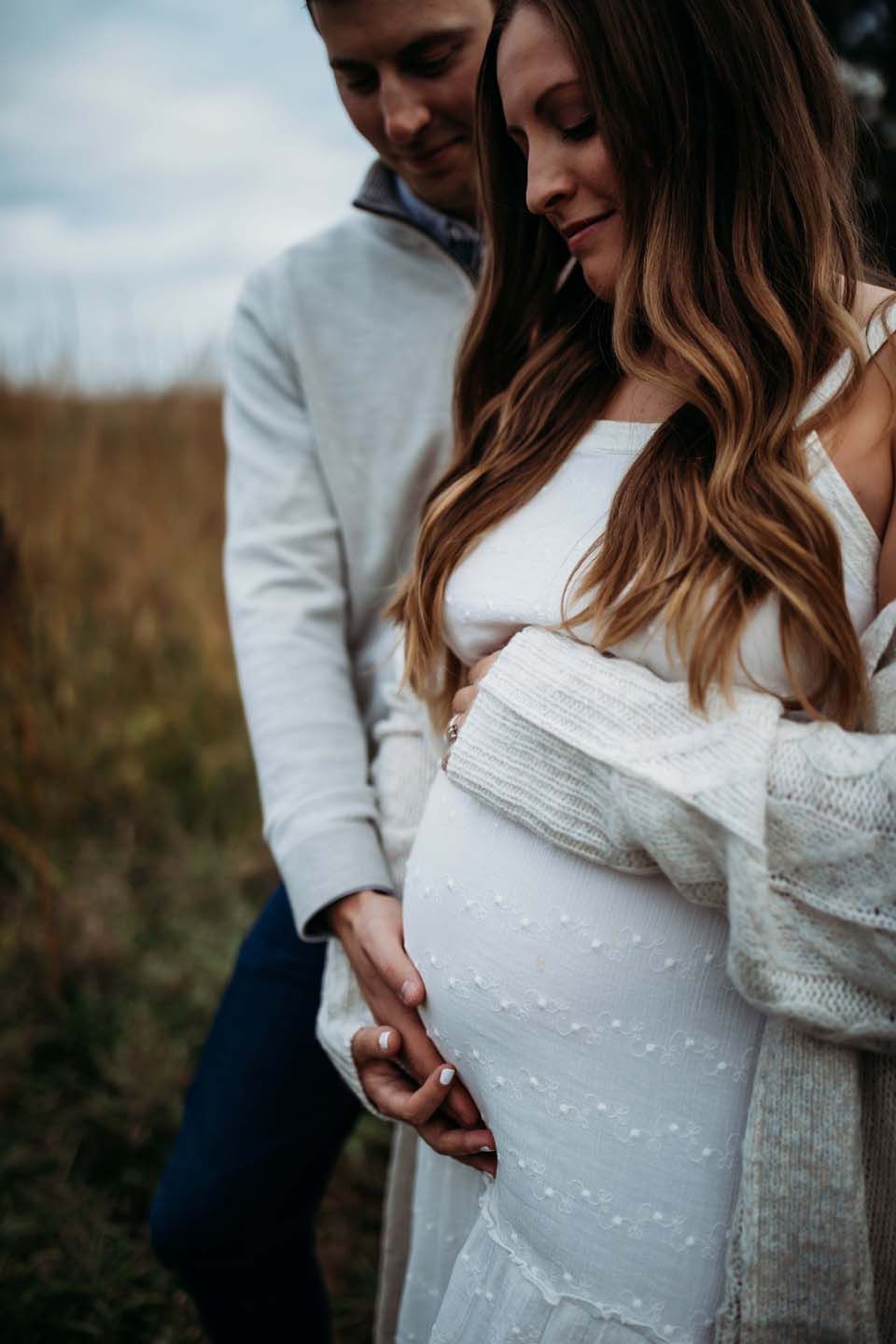 Couple sharing intimate maternity moment outdoors with woman in white dress and grey cardigan against natural backdrop.