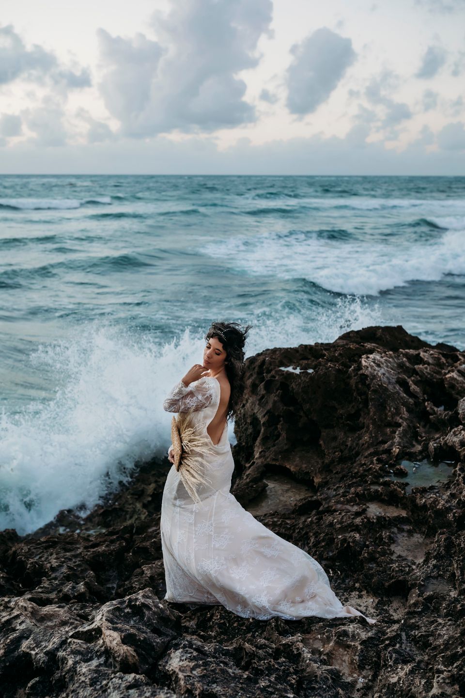 Woman in flowing white dress poses on rocky shore as ocean waves crash behind her.