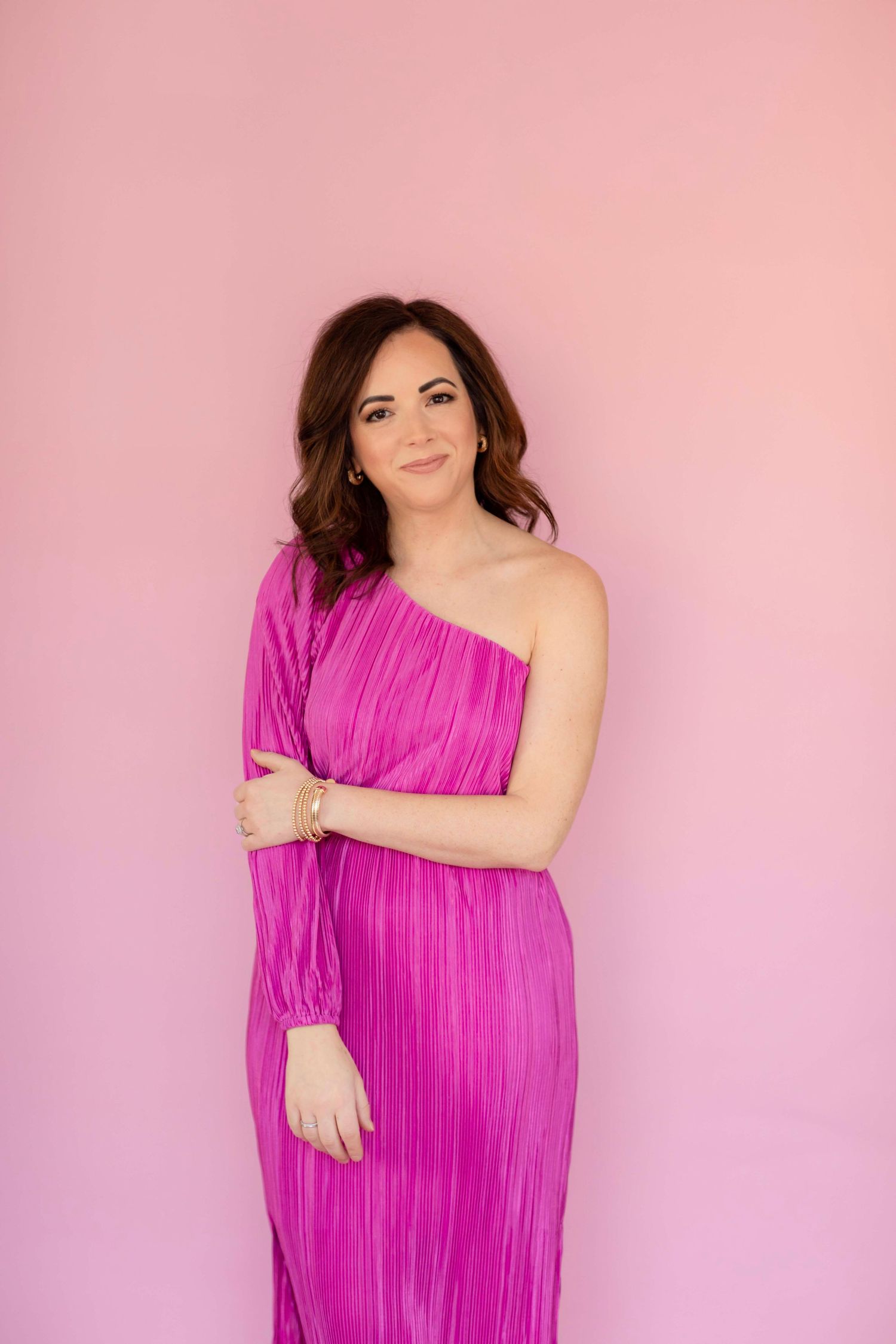 Elegant pose in bright magenta one-shoulder pleated dress against pink gradient backdrop.